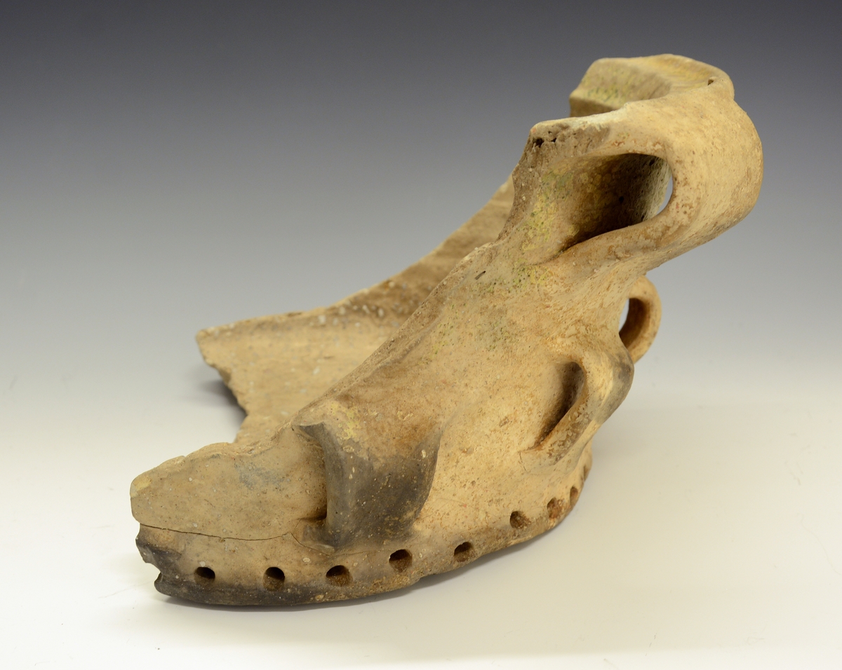 Fra protokollen: 
Keramikkskår. Tykt leirgods, flat bunn, utoverskrånende korpus som ender i en markert brutt kant oppe. Krukka har 4 dekorative ører nede på korpus og (1) og 2 større oppe. En mengde hull går inn i godset i bunnen og fra sidene av bunnen. Krukka mangler glasur, men har partivis (delvis) et slags grønnaktig belegg. Litt over halvparten av bunnen er bevart, men det meste av korpus er slått vekk. Krukka ble funnet i den under BM.1964:198, i merknadene, omtalte Bølebåten.