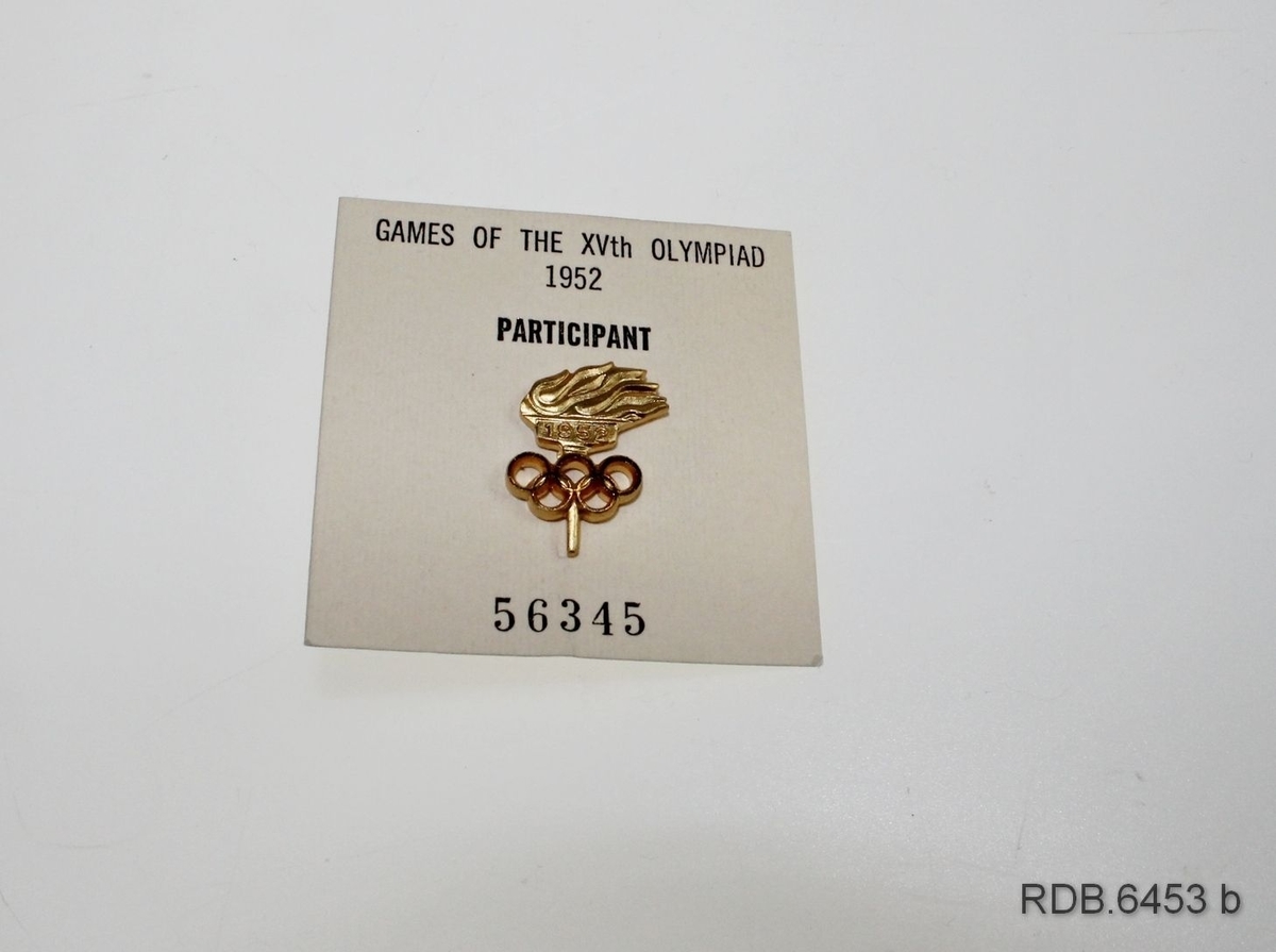 Gullfarget deltagerpin fra OL i Oslo 1952 til å feste på jakkeslaget. Selve pinen forestiller en fakkel med den olympiske ild med de olympiske ringer under. Under flammen står det 1952. Pinen er montert på en papplate med påskrift som ligger i en gjennomsiktig plastlomme. Plastlomma er festet til innsida av et kvitt plastetui som kan brettes sammen og lukkes med en trykknapp.