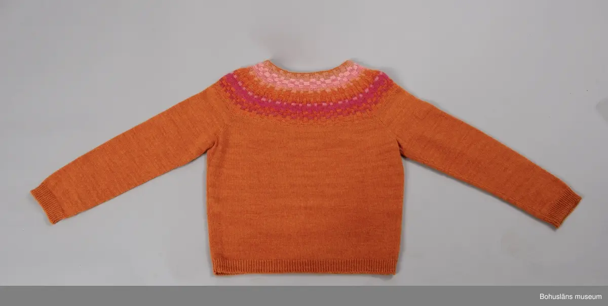 Nyproduktion av stickad jumper med lång ärm, stickad enligt "gamla rundstickningen". Oket är mönsterstickat med räta och aviga maskor i infärgade nyanser av orange och rosa, övriga partier slätstickade i orange. Dubbelvikt halsringning, på baksidan resårstickad. Resårstickade ärmmuddar och nederkant. Fastsydd etikett på insidan halsringningen med vävd text:
"BOHUSLÄNS MUSEUM efter original BOHUS STICKNING".