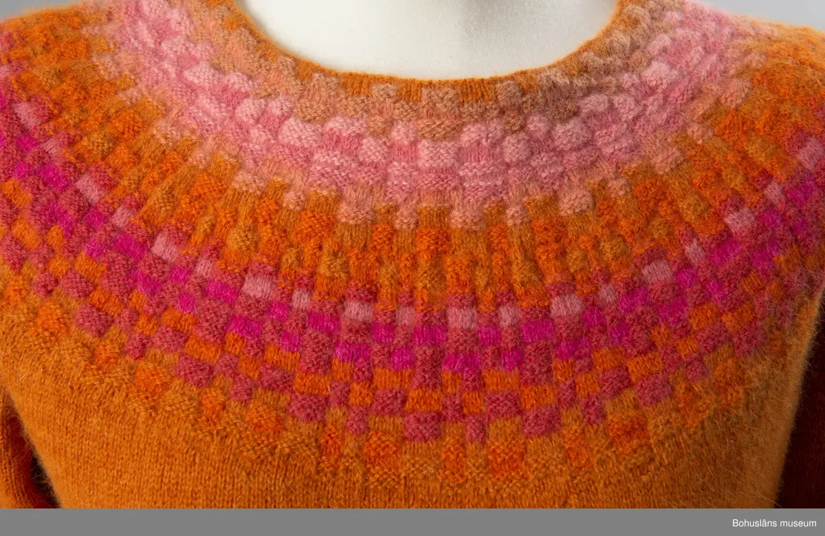 Nyproduktion av stickad jumper med lång ärm, stickad enligt "gamla rundstickningen". Oket är mönsterstickat med räta och aviga maskor i infärgade nyanser av orange och rosa, övriga partier slätstickade i orange. Dubbelvikt halsringning, på baksidan resårstickad. Resårstickade ärmmuddar och nederkant. Fastsydd etikett på insidan halsringningen med vävd text:
"BOHUSLÄNS MUSEUM efter original BOHUS STICKNING".