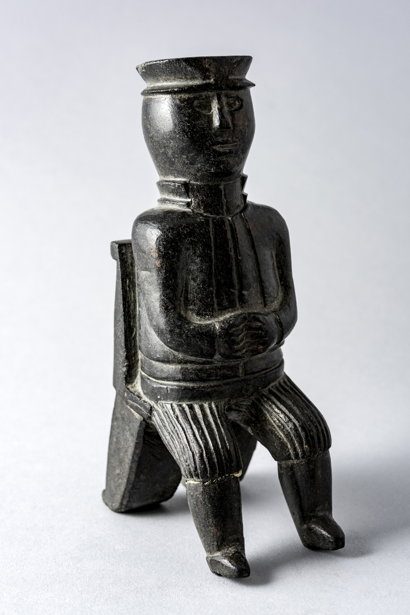 Liten steinskulptur som forestiller mann med hatt sittende på stol uten sete. Bokstøtte?
