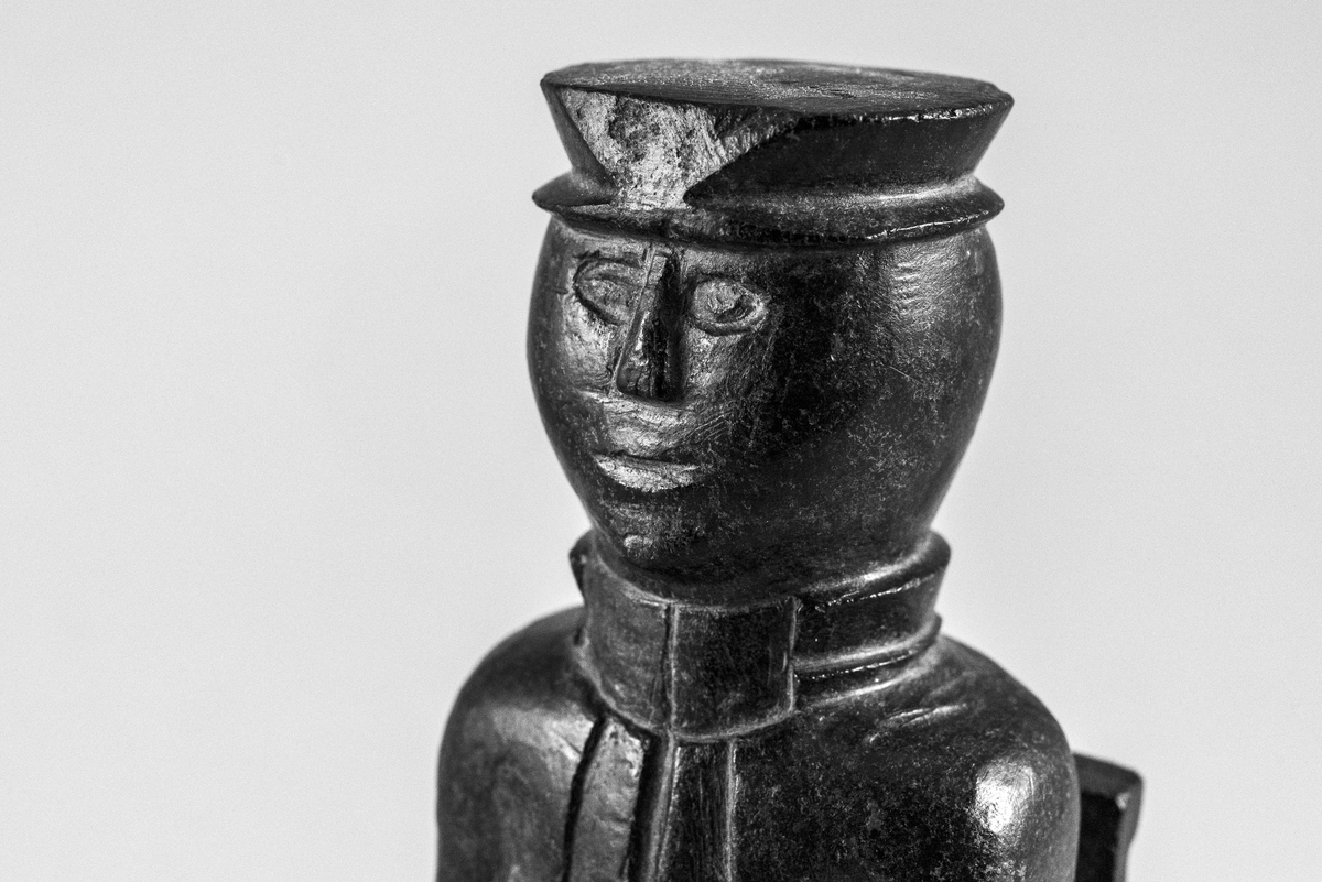 Liten steinskulptur som forestiller mann med hatt sittende på stol uten sete. Bokstøtte?