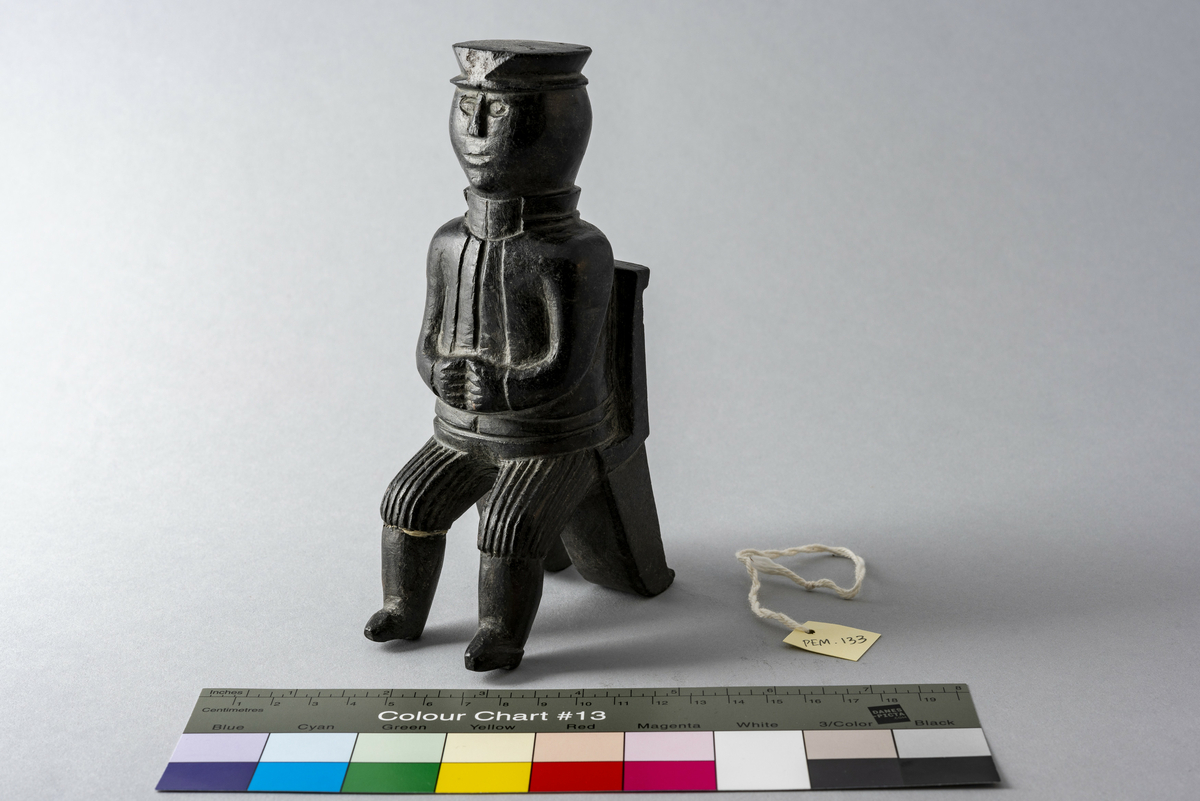 Liten steinskulptur som forestiller mann med hatt sittende på stol uten sete. Bokstøtte?