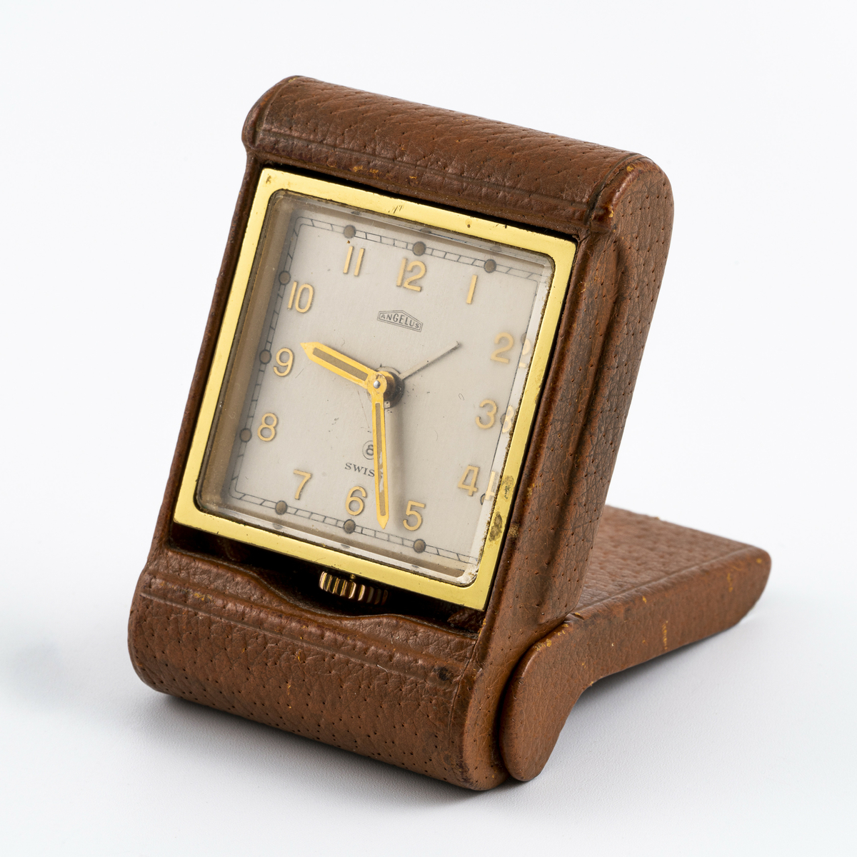 Reiseklokke fra merket Angelus fra Sveits. Ur med i messingfarge festet i brunt skinnetui. Med lokk som i åpen posisjon fungerer som støtte slik at det kan stå på bordet. Uret kan vippes fram for å avdekke baklokk med innstillingsknapper. Har tilhørt Sara Fabricius.