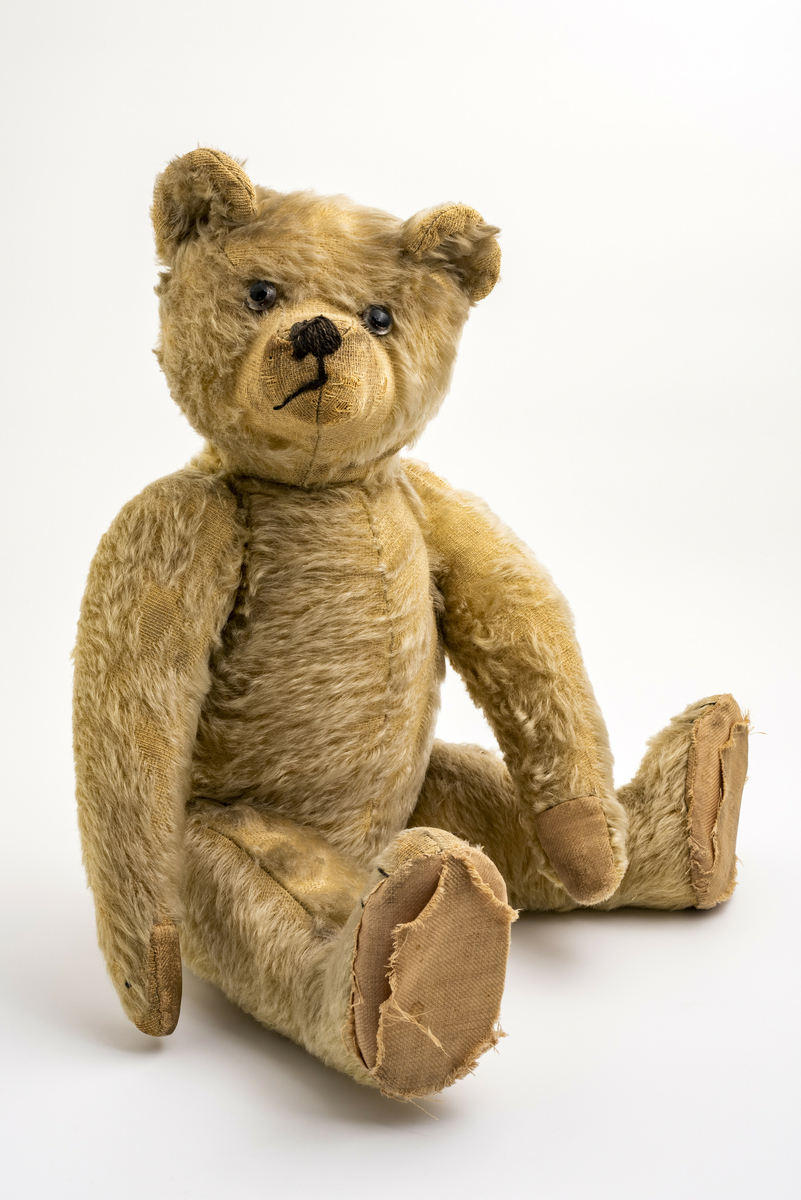Stor teddybjørn med halmfyll (?) og kunstig pels. Alle lender er bevegelige. Påsydd snute, munn og "tær" på potene. Glassøyne. Har tilhørt Sara Fabricius.