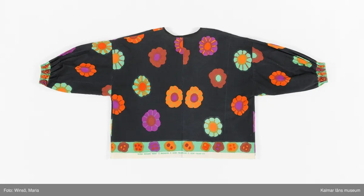 KLM 46157:644. Jacka. För dam. Av svart blommönstrat tryckt tuskaftat bomullstyg. Blommorna är stora, färgglada och strödda över tyget. Rak modell. Utan knäppning, kanter kantade med svart snedslå. Ärm avslutad med insydd resår. Tygets stadkant synlig i jackans nederkant, som en detalj. Tryck i grönt på stadkanten: KATUJI WAKISAKA "NEKKU" © MARIMEKKO OY SUOMI FINLAND 1972.