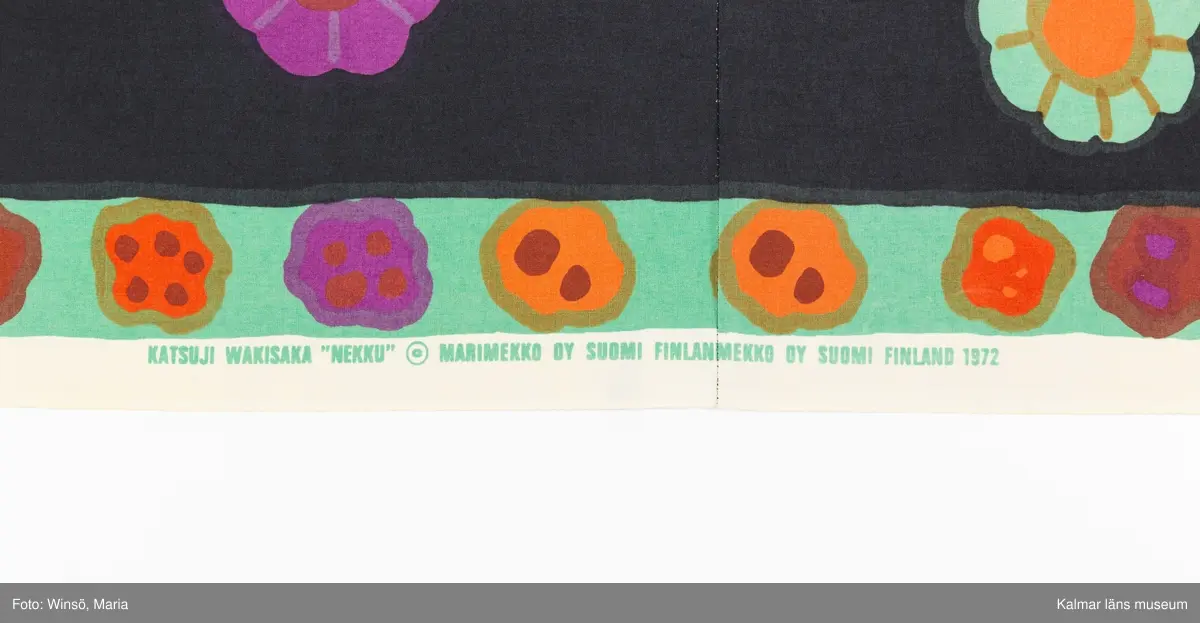 KLM 46157:644. Jacka. För dam. Av svart blommönstrat tryckt tuskaftat bomullstyg. Blommorna är stora, färgglada och strödda över tyget. Rak modell. Utan knäppning, kanter kantade med svart snedslå. Ärm avslutad med insydd resår. Tygets stadkant synlig i jackans nederkant, som en detalj. Tryck i grönt på stadkanten: KATUJI WAKISAKA "NEKKU" © MARIMEKKO OY SUOMI FINLAND 1972.