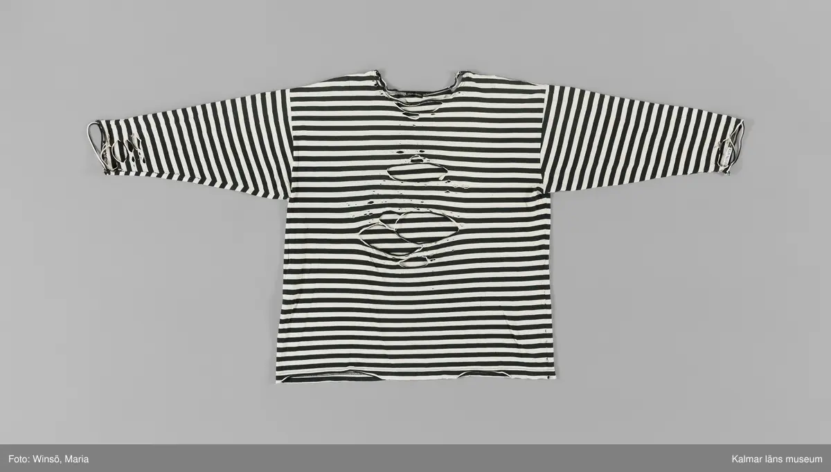 KLM 46157:659. Tröja. Av svart och vit randig bomullstrikå. Rund halsringning. Trekvartslång ärm. Mycket sliten, stora nötta hål på framsidan och ärmarna. Svart textiletikett i nacken med vävd text i vitt: Gudrun Sjödén. Fastsydd etikett med material och tvättråd på insidan i höger sidsöm. 100% bomull, tvättas i 40 grader, strykes med två prickar samt ej torktumling.