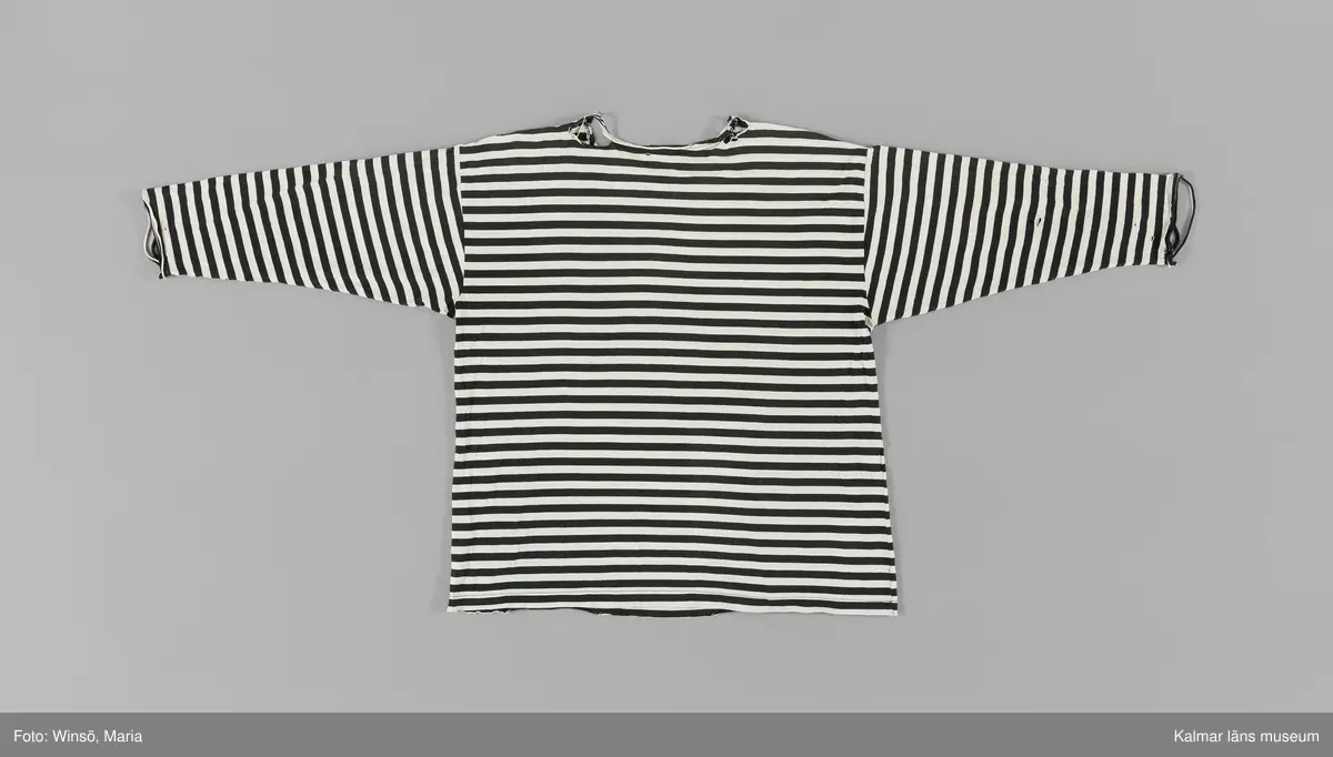 KLM 46157:659. Tröja. Av svart och vit randig bomullstrikå. Rund halsringning. Trekvartslång ärm. Mycket sliten, stora nötta hål på framsidan och ärmarna. Svart textiletikett i nacken med vävd text i vitt: Gudrun Sjödén. Fastsydd etikett med material och tvättråd på insidan i höger sidsöm. 100% bomull, tvättas i 40 grader, strykes med två prickar samt ej torktumling.