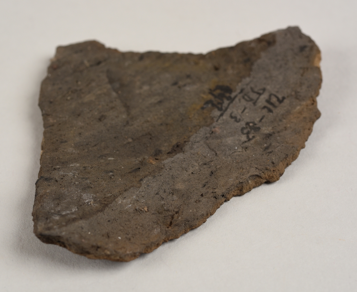 Lett buet keramikkfragment med bruddkanter på alle sidene. Fragmentet er svart i farge på utsiden og grått i farge på innsiden. Bruddkantene er grå i farge.