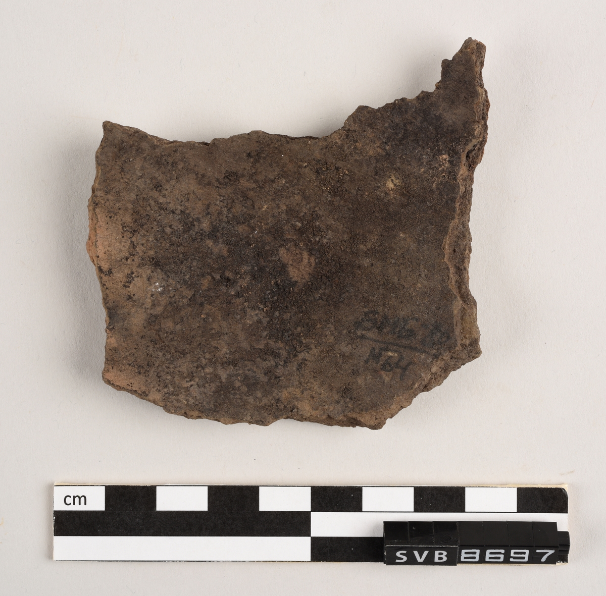 Lett buet keramikkfragment med bruddkanter på alle sidene. Fragmentet er brunt i farge. Bruddkantene er brune i farge.