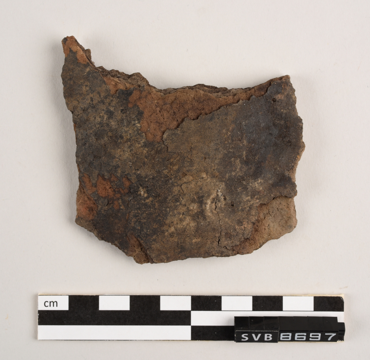 Lett buet keramikkfragment med bruddkanter på alle sidene. Fragmentet er brunt i farge. Bruddkantene er brune i farge.