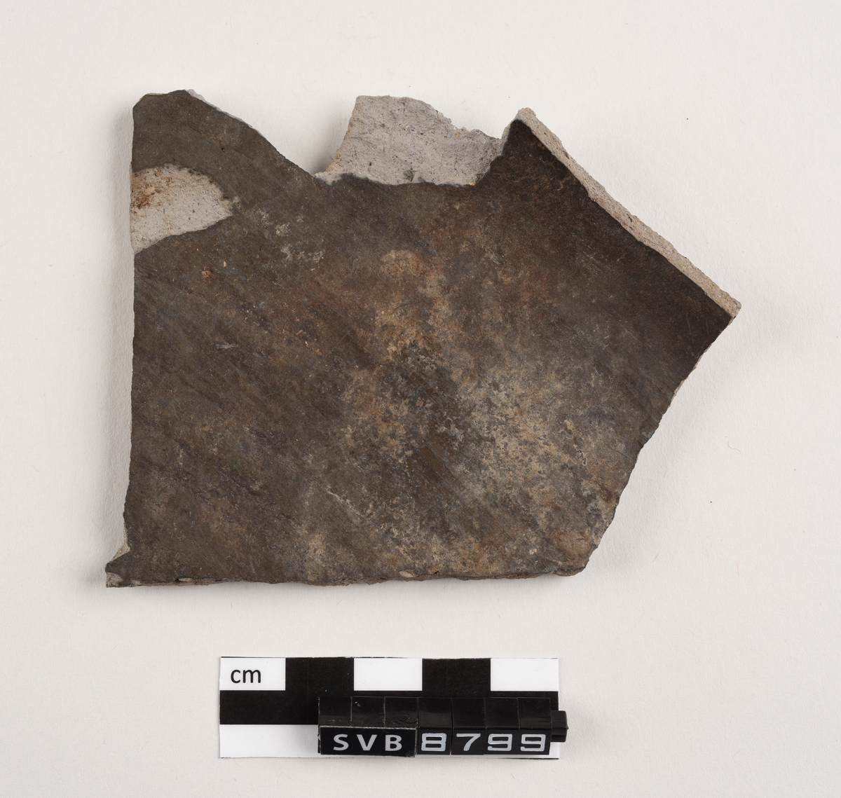 Flatt keramikkfragment med bruddkanter på alle sidene. Fragmentet er glasert på den ene siden. Fragmentet og bruddkantene er grå i farge.