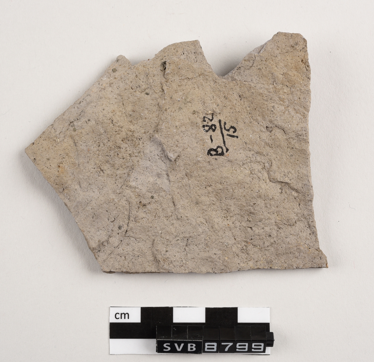 Flatt keramikkfragment med bruddkanter på alle sidene. Fragmentet er glasert på den ene siden. Fragmentet og bruddkantene er grå i farge.