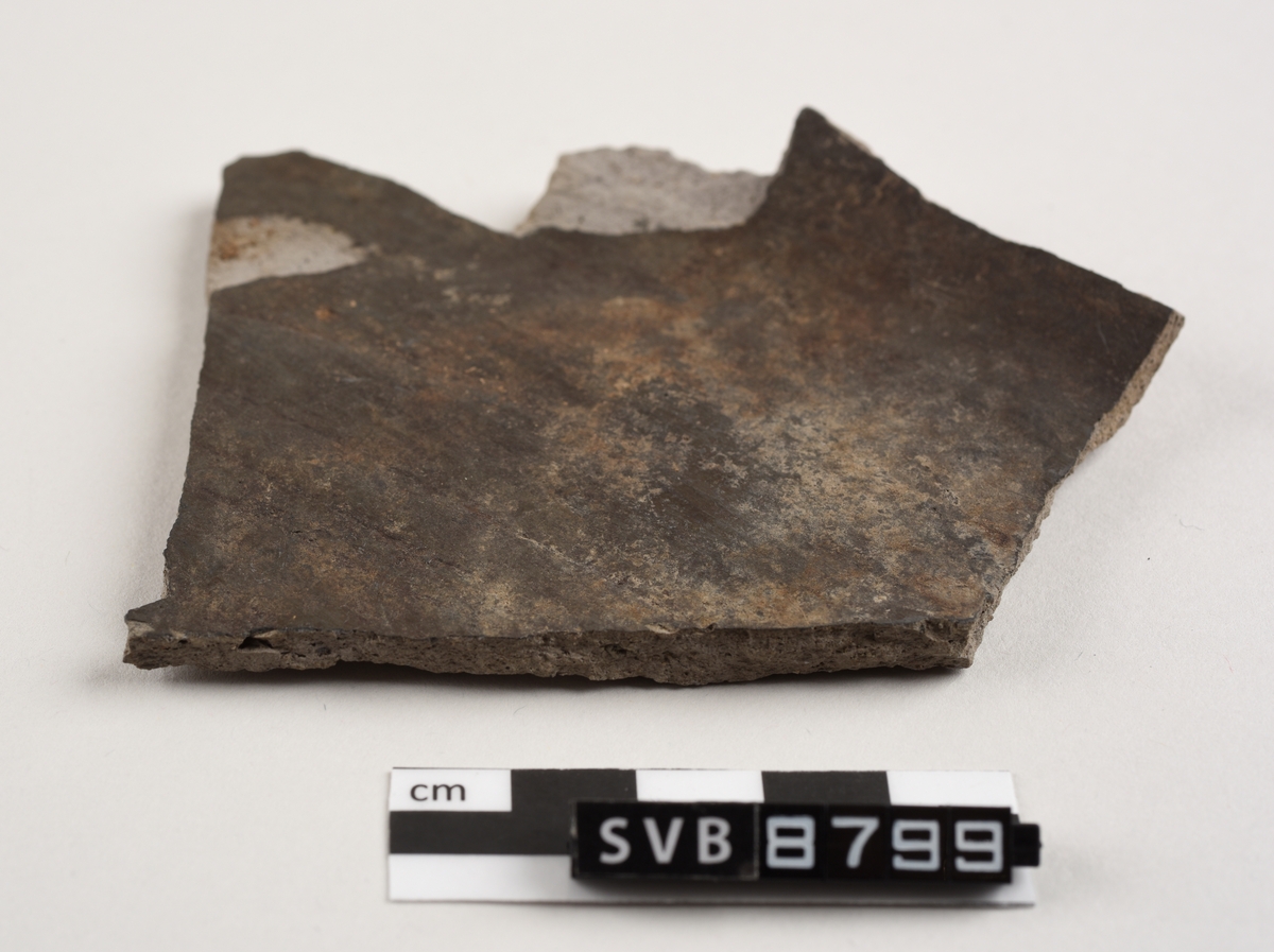 Flatt keramikkfragment med bruddkanter på alle sidene. Fragmentet er glasert på den ene siden. Fragmentet og bruddkantene er grå i farge.