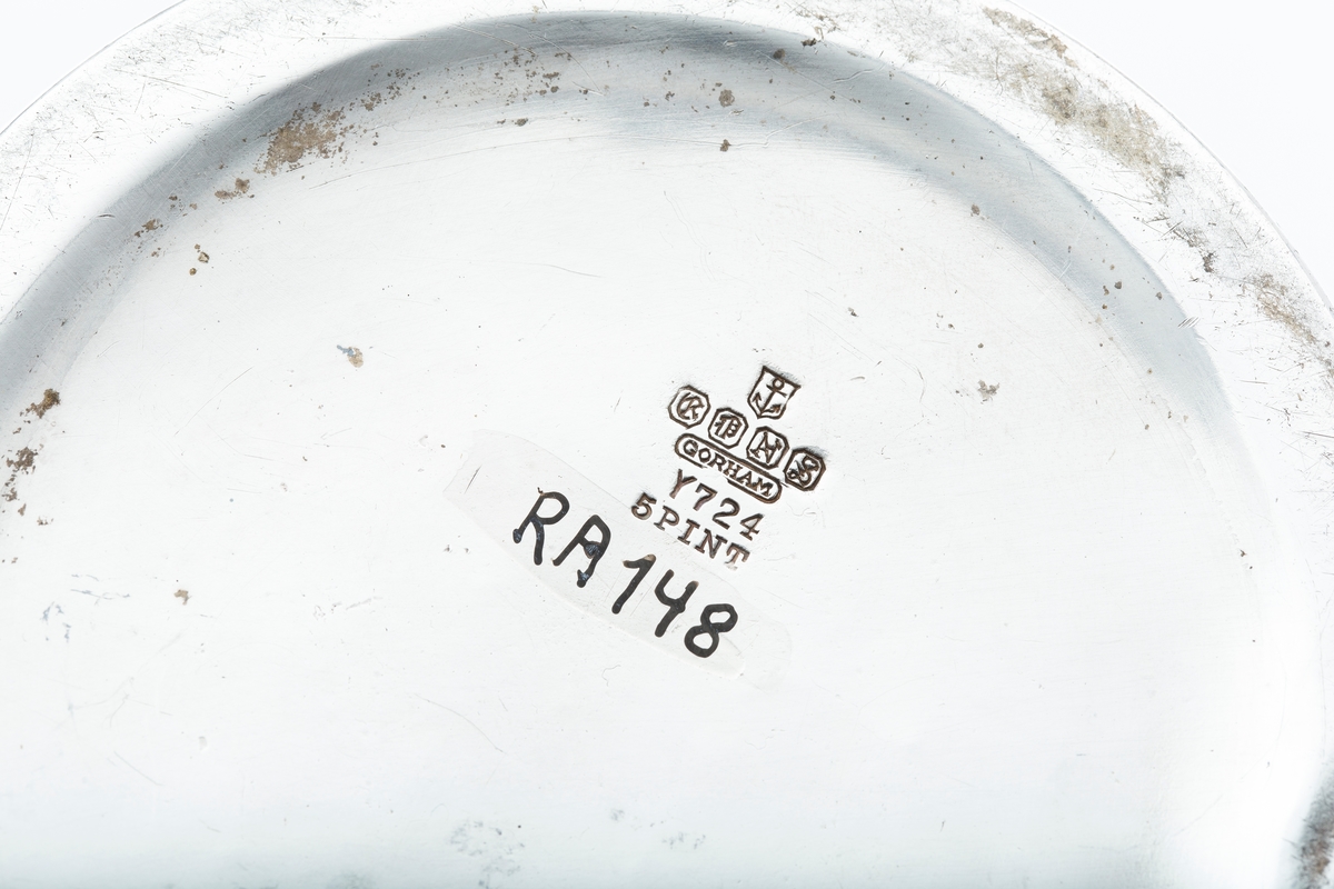 Mugge, blankt metall, bred tut, slank hank. Smal bord rundt muggen med høyt relieff, derunder siselerte blomsterrosetter, bånd og medaljonger. Stempel:? Under bunden er innrisset JVB. 