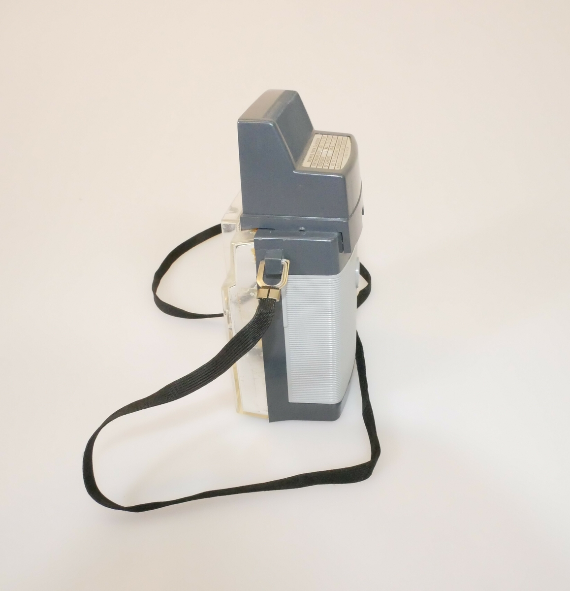 Rektangulært fotoapparat. Lysegrå forside dekket av klar hard plast, midt på er det en linse. På toppen er det boks av grå hard plast til blits. På baksiden er det grå og lysegrå hard plast. Sort bånd festet på begge sider av kameraet med metallfeste. 