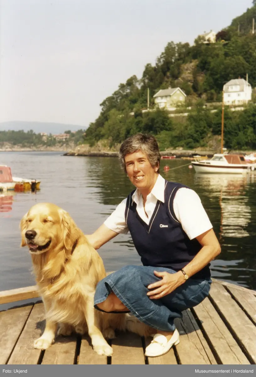 Kvinne i marineblå vest, saman med hund på ei brygge.