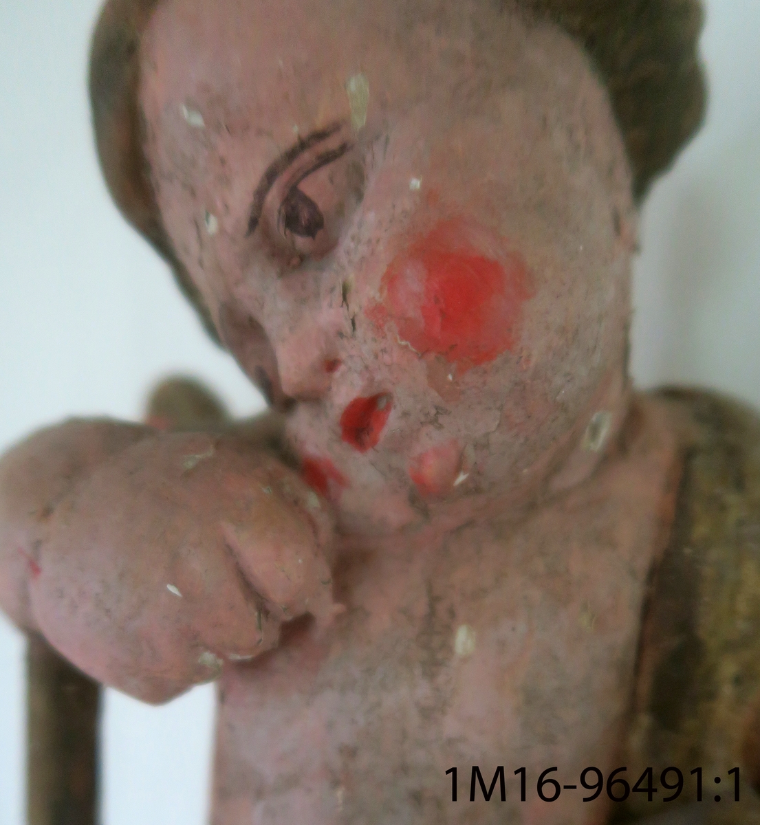 Skulpterad ängel av trä. Sitter på dödskalle med huvudet något högerböjt. Håller en snäcka med tre frukter (?) i höger hand. Draperat tygstycke runt midjan, över höger axel och rygg, avslutas framtill. Saknar vingar, utsträckningar i skuldrorna visar var de suttit. Vänster arm och höger fot saknas. Har hållit ett nu avbrutet föremål i munnen. Träplugg i botten avbruten. Sekundär målning i mörk hudfärg med röda kinder. Hår, dödskalle och övriga detaljer i brunt. Färgen fläckvis borta. Har troligen tillhört epitafium eller altaruppsats.