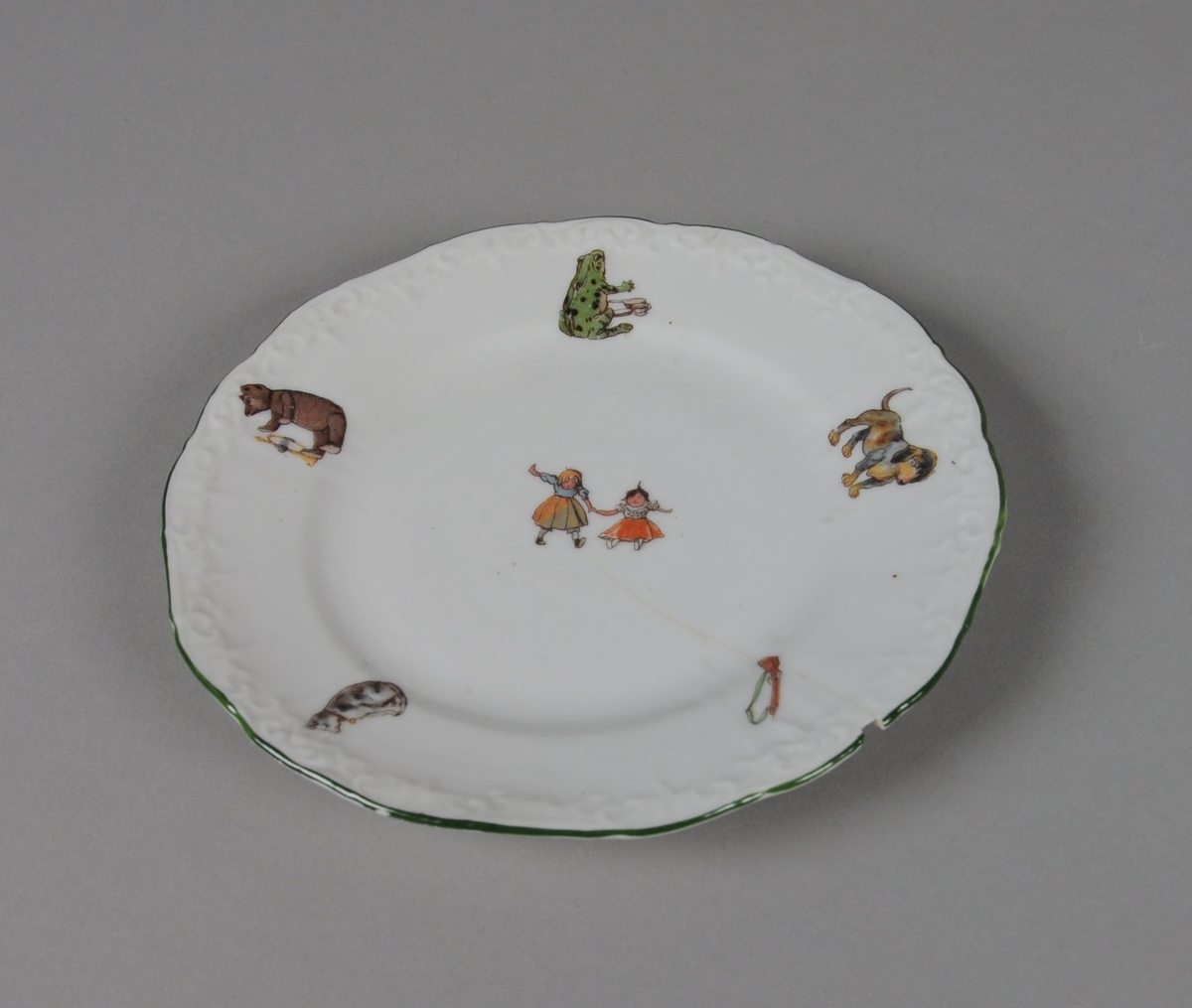 Seks leketallerkener av glassert keramikk. Randen er bølgete og har en dekorativ grønn stripe. Flerfarget trykk med motiv av dyr og leker, hver tallerken har forskjellige kombinasjoner av trykkene.