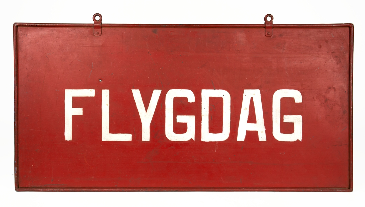 Skylt av plåt med två öglor upptill för uppsättning. Skylten är målad röd med vit text på båda sidor. Text ena sidan: FLYGDAG. Text andra sidan: ICKE FLYGDAG. I plåten finns en skada i form av ett mindre hål.