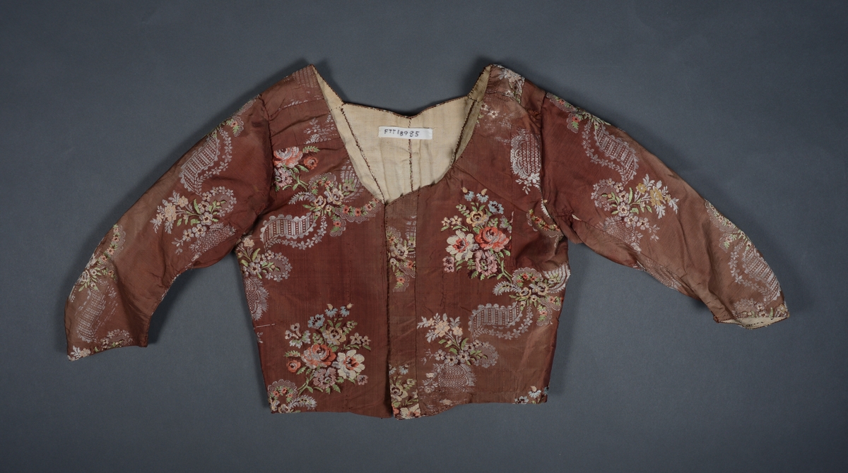 Et kjoleliv/jakke med lange ermer sydd av brun silke. I silken er det ivevd blomstermønster i rødt, grønt, brunt, grått, rosa og gult. Mellom blomstermotivene er det ivevd grått "blondemønster". Kjolelivet er foret med naturfarget lin. I front er det 7 par hull for snøring. Snøringen dekkes av en langsgående påsydd klaff av brun silke med blomstermotiv. Midt bak er det sydd i en kile for form. Kjolelivet er oppstivet med 11 spiler.