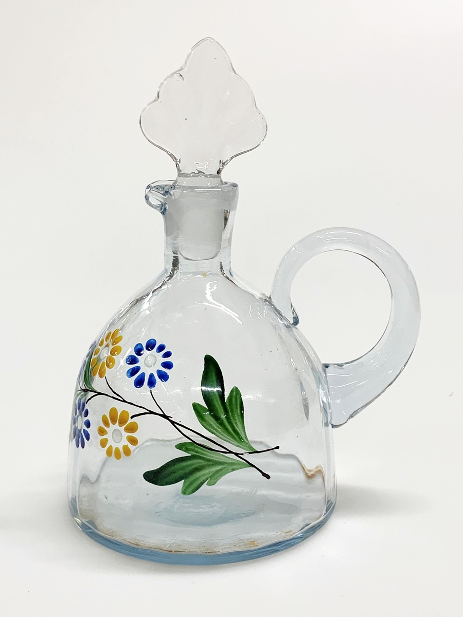 Mindre blåtonad karaff (sherry eller likör) med optikblåst och målad kropp som övergår i en kort hals med pip. Pressad propp. Det målade motivet består av en blomsterkvist med blå och gula blommor.
