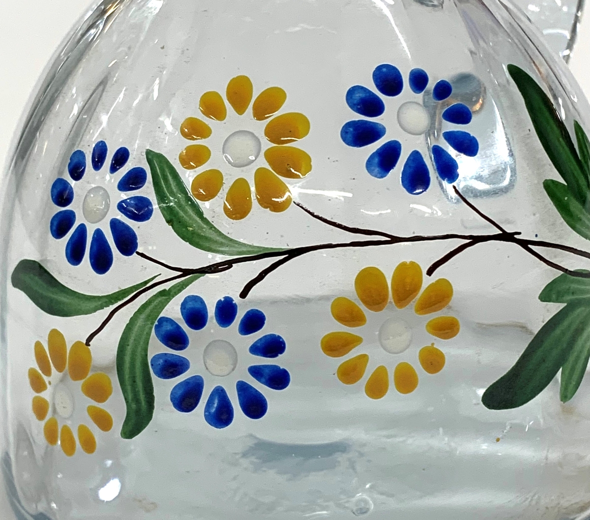 Mindre blåtonad karaff (sherry eller likör) med optikblåst och målad kropp som övergår i en kort hals med pip. Pressad propp. Det målade motivet består av en blomsterkvist med blå och gula blommor.
