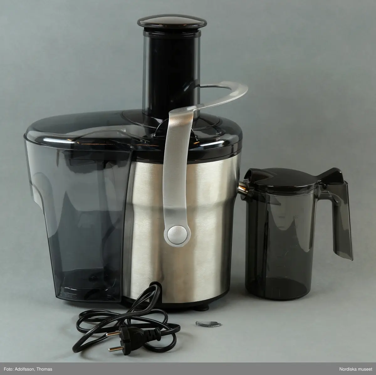 En elektrisk råsaftcentrifug av märket Elektrolux PERFECTJUICE ESF 2000. Består av  6 delar. a) Själva råsaftcentrifugen är cylinderformad och av metall och plast. De övriga delarna är av rökgrå hårdplast och består av b+) ett lock med c) press samt en d) behålllare som fruktköttet samlas i och e) en liten lockförsedd juicekanna med f) skumseparator i form av en plasskena som saften rinner ut i när den är klar. Dessutom en g) bruksanvisningsbroschyr och ett h) garantipapper.
Locket (b+) har en skada där en bit är avslagen.

Lena Kättström Höök 2014-01-10