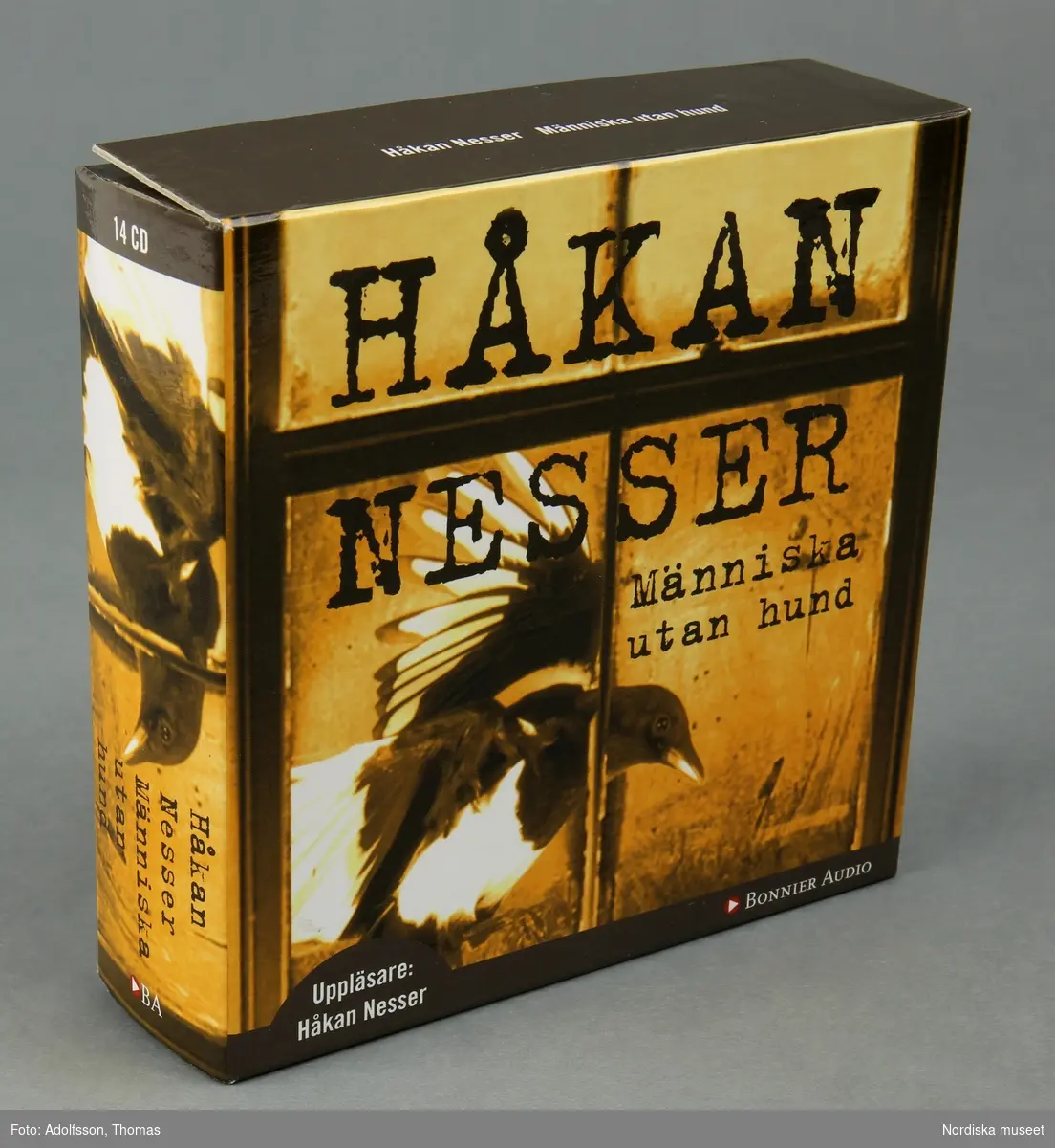 CD-bok.  a:1-2-n:1-2) 14 st CD-skivor, i en kvadratisk, transparent plastficka, innehållandes uppläsning av romanen "Människa utan hund" skriven av författaren Håkan Nesser som även är uppläsaren på denna CD-inspelning.  Inspelningen är totalt 15 timmar lång.

CD-skivorna är tunna cirkelfornade,  12 cm i diameter, på ena sidan oranga med text om titel samt kort om produktionen. Varje skiva är numrerad för att visa rangordningen i vilken skivorna ska lyssnas på/spelas. Baksidan på skivan är silverfärgad, speglliknande, och innehåller själva ljudspåren som läses av när skivan placeras i en CD-spelare.
Varje skiva är separat plackad i en kvadratisk, transparent, plastficka för att skydda mot repor som ev skulle kunna skada skivan. 
Samtliga skivor är tillsammans packade i en o) kvadratisk papperskartong/ konvolut. Konvolutet har på framsidan en bild av ett fönster med ett gultonat fotografi visandes ett fönster med en flaxande fågel utanför. På konvolutets baksida finns en introduktion till bokens handling samt  information om tillverkningen av denna CD-bok.
(CD är en förkortning av engelskans Compact Disc. CD-skivan kan lagra digital information, exempelvis musik.
CD skivans konstruktion: överst är ett transparant plastlager följt av ett tunt lager av blank metall med ett präglat spiralformat spår med mikroskopiskt små gropar som avläses av CD-spelarens laserstråle när skivan snurrar (ca 500-700 varv/minuten). Ljuset studsar tillbaka och läses som en följd av ettor och nollor, som i detta fall, blir till ljud.(Källa/. Nationalencyklopedin)


/Cecilia Wallquist 2019-02-20