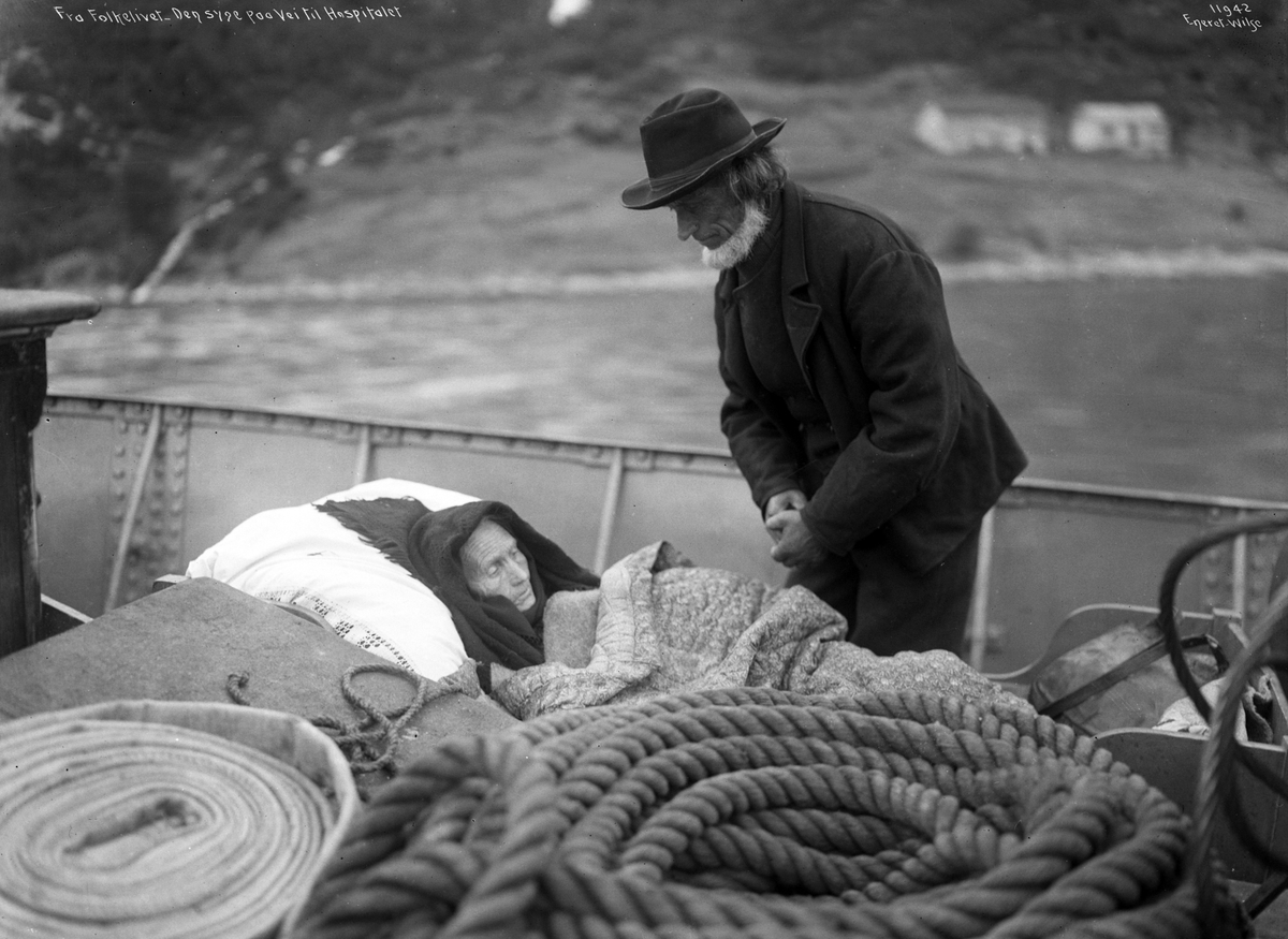 Syketransport av et eldre par i båt på vei til Ålesund. "Den syke paa Dekket". Ingeborg og Ola Hustad, Hjørundfjord.