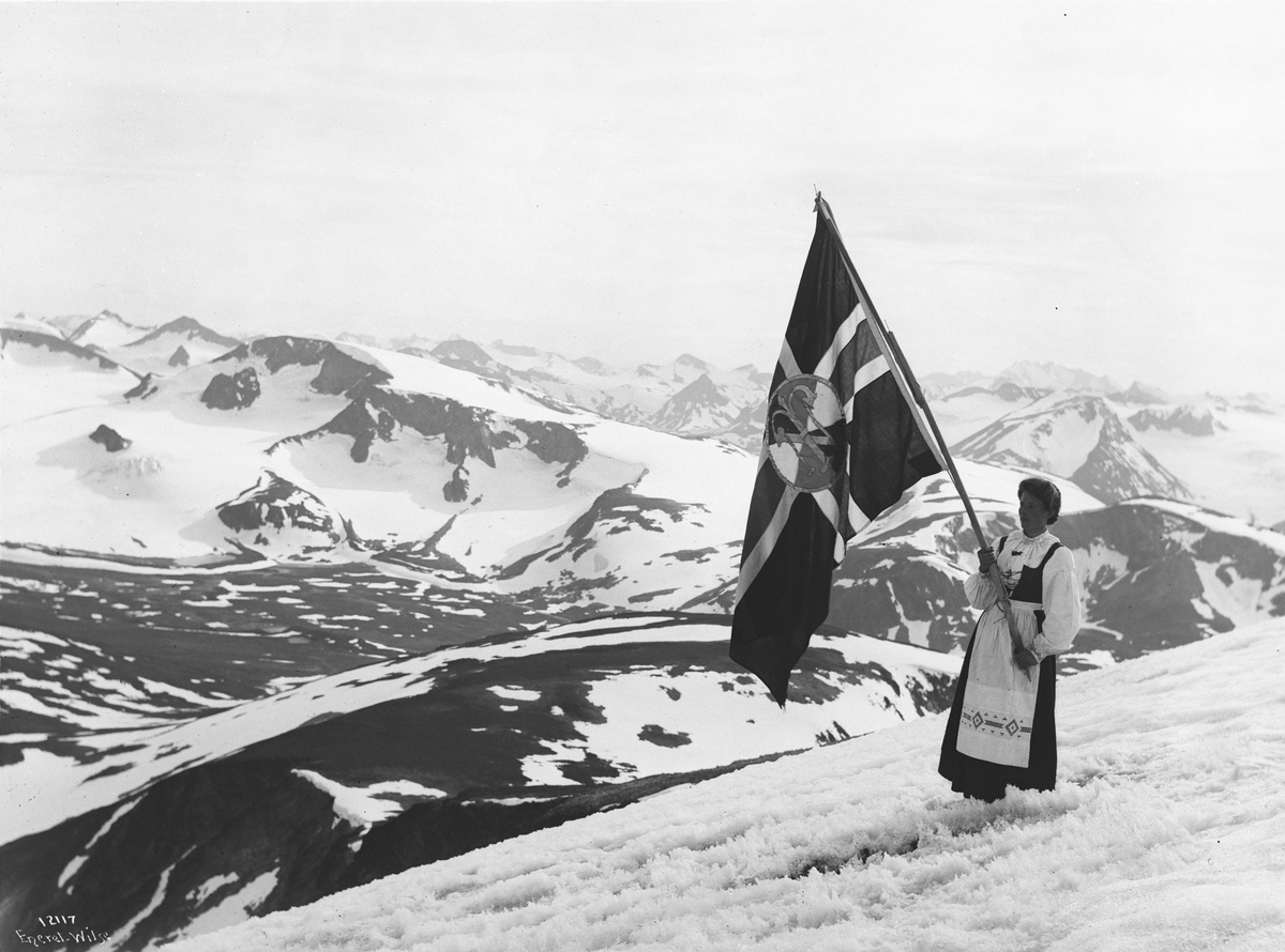 Bunadkledd kvinne, Hanna Bekkevoll, med Turistforeningens Flagg. Antatt på Glittertind, Jotunheimen Nasjonalpark, Lom, Innlandet (Oppland).