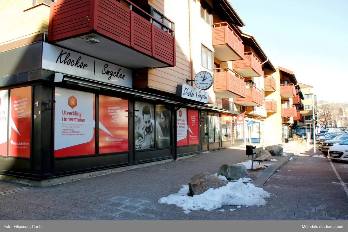 Hörnet på huset med adressen Bergmansgatan 6 i Mölndals Centrum. Bostäder ovanför entré och skyltfönster till F.M. Ericsson Ur & Guldsmide AB, Salong Hårfager och Direkten Mölndal Cigarr & Tobak i gatuplanet. Fotografi taget den 26 februari 2018. Byggnadsdokumentation inför rivning.