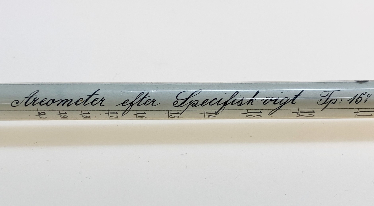 Areometer med tillhörande papprör, användes för att bestämma specifik vikt på vätskor. Påminner om en termometer. Nederst en uppblåst kula med flytande kvicksilver, vilken övergår i ett smalt rör som i sin tur övergår i ytterligare en (tom) kula. I "mätdelen" ett papper med skala och texten: "Areometer efter Specifisk vigt Tp: 15 C".