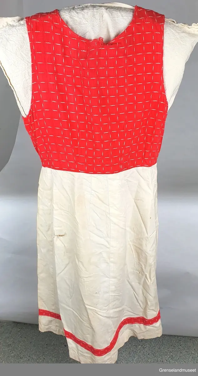 "Hverdagsbunad"  Bomull. 1955.60. Håndsydde huller til snøring i front.
Rødt mønstret liv. Hvitt skjørt med kant av det røde stoffet.