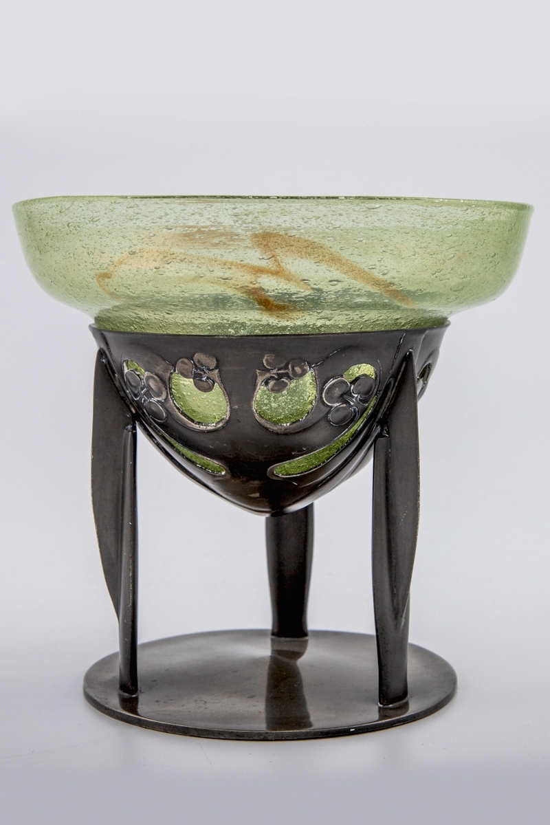 Glasskål som hviler på trebent tinnfot.  Skålen er klokkeformet med bred kant, og den eplegrønne glassmassen er fylt med små blærer. Langs kanten er det partier med innlagt bladgull. Den nedre delen bæres av en trebent fot i tinn, hvis overflate er oksidert. Den øvre delen er gjennombrutt, og smykket med stiliserte planteornamenter i lavt relieff. De tre benene hviler på sirkulær fotplate, som er noe hevet mot midten.