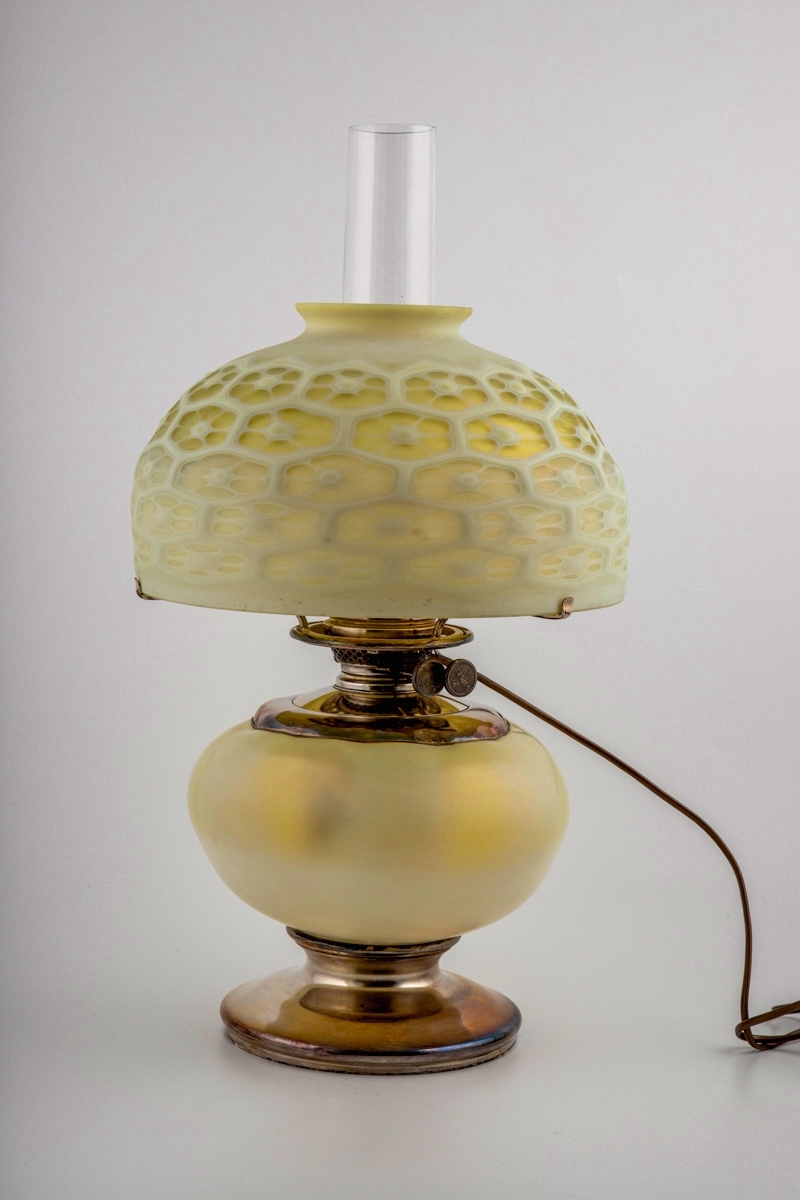 Bordlampe for parafin i farget glass. Lampen hviler på en sirkulær fot i forsølvet bronse. Oljehuset er plassert i lampens nedre del, og er innkapslet i en ovalformet kule av opakt glass med et grønnlig skjær. Brenneren og skruen – sistnevnte for å justere veken – er også utført i forsølvet bronse. Det sylindriske lampeglasset, hvis nedre er kuleformet, er delvis skjult under en bred halvkuleformet skjerm. Skjermens ytterside er dekorert med et bikubelignende mønster i lavt relieff, hvor fordypningene har en grønnlig farge, mens de opphøyde partiene er melkehvite.