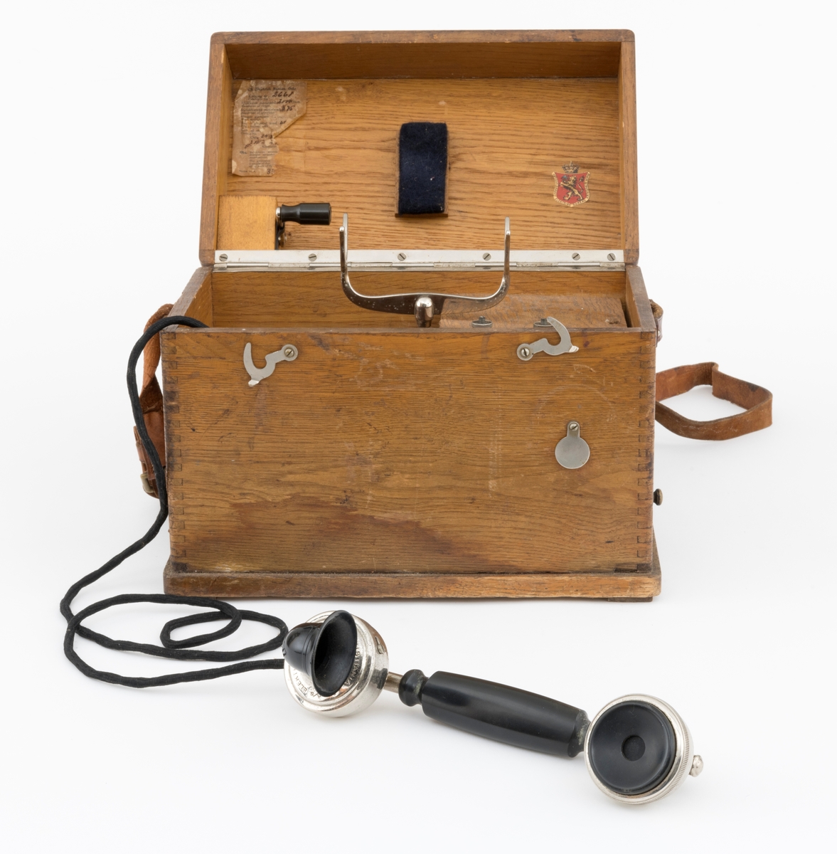 Bærbar felttelefon, antakelig av type som ble brukt tidlig på 1900-tallet. Telefonen er montert i ei trekasse, antakelig lagd av eikematerialer. Kassa har ei botnplate som er 33,1 centimeter lang, 20,2 centimeter bred og 1,5 centimeter tjukk. Ved endene av botnplatas underside er det 19,3 centimeter lange og 3,1 centimeter brede trelister som  gjenstanden sto på. Oppå den nevnte botnplata er det ei 32 centimeter lang, 19 centimeter bred og 16,6 centimeter høy kasse med fingerskjøtte, noe avrundete hjørner. Til denne kassas bakre langside er det hengslet et lokk av samme type materiale og med samme lengde og bredde som kassa. Lokket er 6,1 centimeter høyt. Lokket stenges ved hjelp av dreibare kroker på kassas frontside som vendes, slik at de omslutter skruer på lokkets frontside. Litt lengre neden på kassefronten er det et gjennombrutt hull med 1,3 centimeters diameter, antakelig innføringspunktet for sveiva til telefonen. Dette hukket kan stenges ved hjelp av ei lita, dreibar stålskive når sveiva ikke er i bruk. Da festes sveiva i en trekloss som er innlimt i et hjørne i lokket. Telefonen bæres i ei cirka 2,4 centimeter bred lysebrun lærrein, som er festet i messingknapper, sentralt på den nedre delen av kassas tverrender. At kassa bæres med den riktige sida opp er sikret ved hjelp av smøyestoler av lær høyere oppe på de samme endeflatene. Lengden på bærereima kan justeres ved hjelp av ei nålespenne. Inne i kassa er det antakelig et batteri, skjult under trelokk i to plan. På det nedre er det inntappet en toleddet, forkrommet holder for telefonrøret. Også telefonrøret er forkrommet, med har bakelittkomponenter for håndgrep (midten), øreanlegg og taletrakt. Sentralt på lokkets underside er det pålimt en trekloss med et tjukt svart filtbelegg, som skulle holde telefonrøret på plass under transport. Ellers er det innlimt to etiketter på undersida av lokket. Den ene er et firmamerke som refererer til «AKTIESELSKABET ELEKTISK BUREAU, KRISTIANIA, NORGE». På dette merket er det et rødt skjold med en løvefigur med krone over. Undersida av lokket er det også påklistrert en etikett med tekniske spesifikasjoner fra produsenten. Denne kontrollseddelen har også vært nummert, datert og signert, men hjørnet hvor blant annet årstallet har stått, er dessverre avrevet.