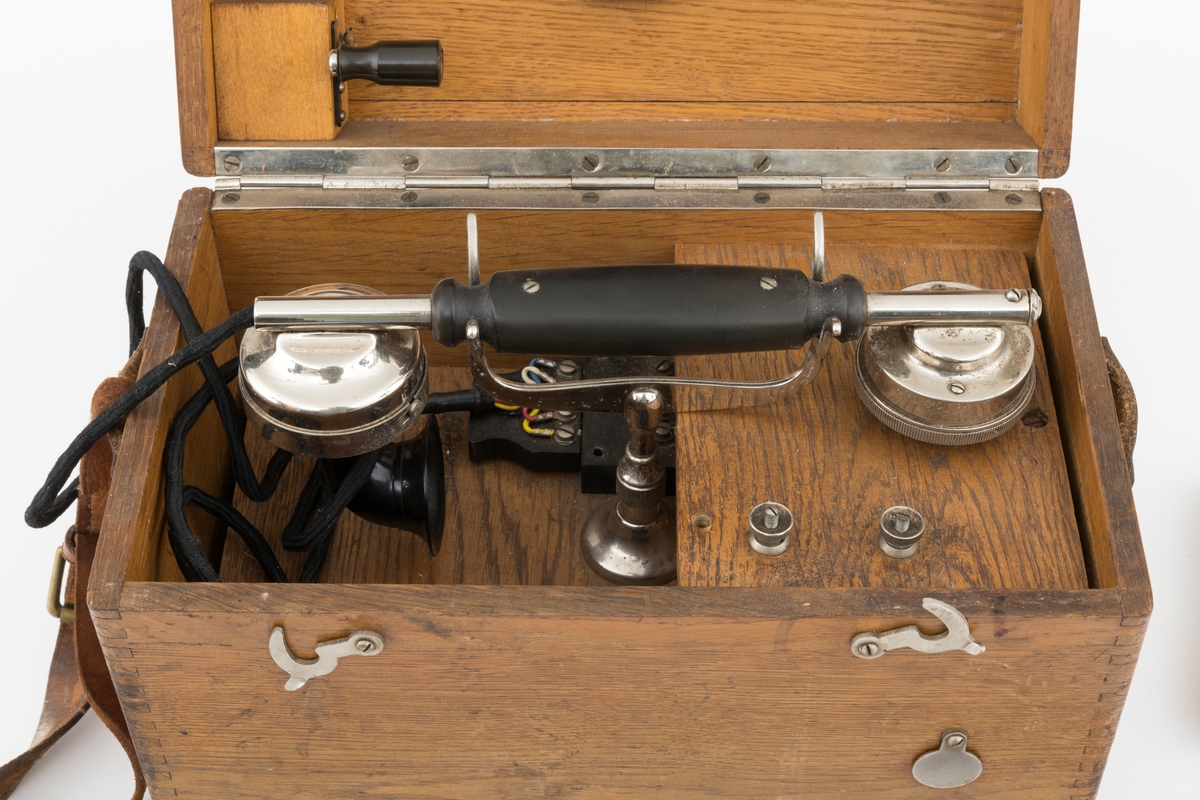 Bærbar felttelefon, antakelig av type som ble brukt tidlig på 1900-tallet. Telefonen er montert i ei trekasse, antakelig lagd av eikematerialer. Kassa har ei botnplate som er 33,1 centimeter lang, 20,2 centimeter bred og 1,5 centimeter tjukk. Ved endene av botnplatas underside er det 19,3 centimeter lange og 3,1 centimeter brede trelister som  gjenstanden sto på. Oppå den nevnte botnplata er det ei 32 centimeter lang, 19 centimeter bred og 16,6 centimeter høy kasse med fingerskjøtte, noe avrundete hjørner. Til denne kassas bakre langside er det hengslet et lokk av samme type materiale og med samme lengde og bredde som kassa. Lokket er 6,1 centimeter høyt. Lokket stenges ved hjelp av dreibare kroker på kassas frontside som vendes, slik at de omslutter skruer på lokkets frontside. Litt lengre neden på kassefronten er det et gjennombrutt hull med 1,3 centimeters diameter, antakelig innføringspunktet for sveiva til telefonen. Dette hukket kan stenges ved hjelp av ei lita, dreibar stålskive når sveiva ikke er i bruk. Da festes sveiva i en trekloss som er innlimt i et hjørne i lokket. Telefonen bæres i ei cirka 2,4 centimeter bred lysebrun lærrein, som er festet i messingknapper, sentralt på den nedre delen av kassas tverrender. At kassa bæres med den riktige sida opp er sikret ved hjelp av smøyestoler av lær høyere oppe på de samme endeflatene. Lengden på bærereima kan justeres ved hjelp av ei nålespenne. Inne i kassa er det antakelig et batteri, skjult under trelokk i to plan. På det nedre er det inntappet en toleddet, forkrommet holder for telefonrøret. Også telefonrøret er forkrommet, med har bakelittkomponenter for håndgrep (midten), øreanlegg og taletrakt. Sentralt på lokkets underside er det pålimt en trekloss med et tjukt svart filtbelegg, som skulle holde telefonrøret på plass under transport. Ellers er det innlimt to etiketter på undersida av lokket. Den ene er et firmamerke som refererer til «AKTIESELSKABET ELEKTISK BUREAU, KRISTIANIA, NORGE». På dette merket er det et rødt skjold med en løvefigur med krone over. Undersida av lokket er det også påklistrert en etikett med tekniske spesifikasjoner fra produsenten. Denne kontrollseddelen har også vært nummert, datert og signert, men hjørnet hvor blant annet årstallet har stått, er dessverre avrevet.