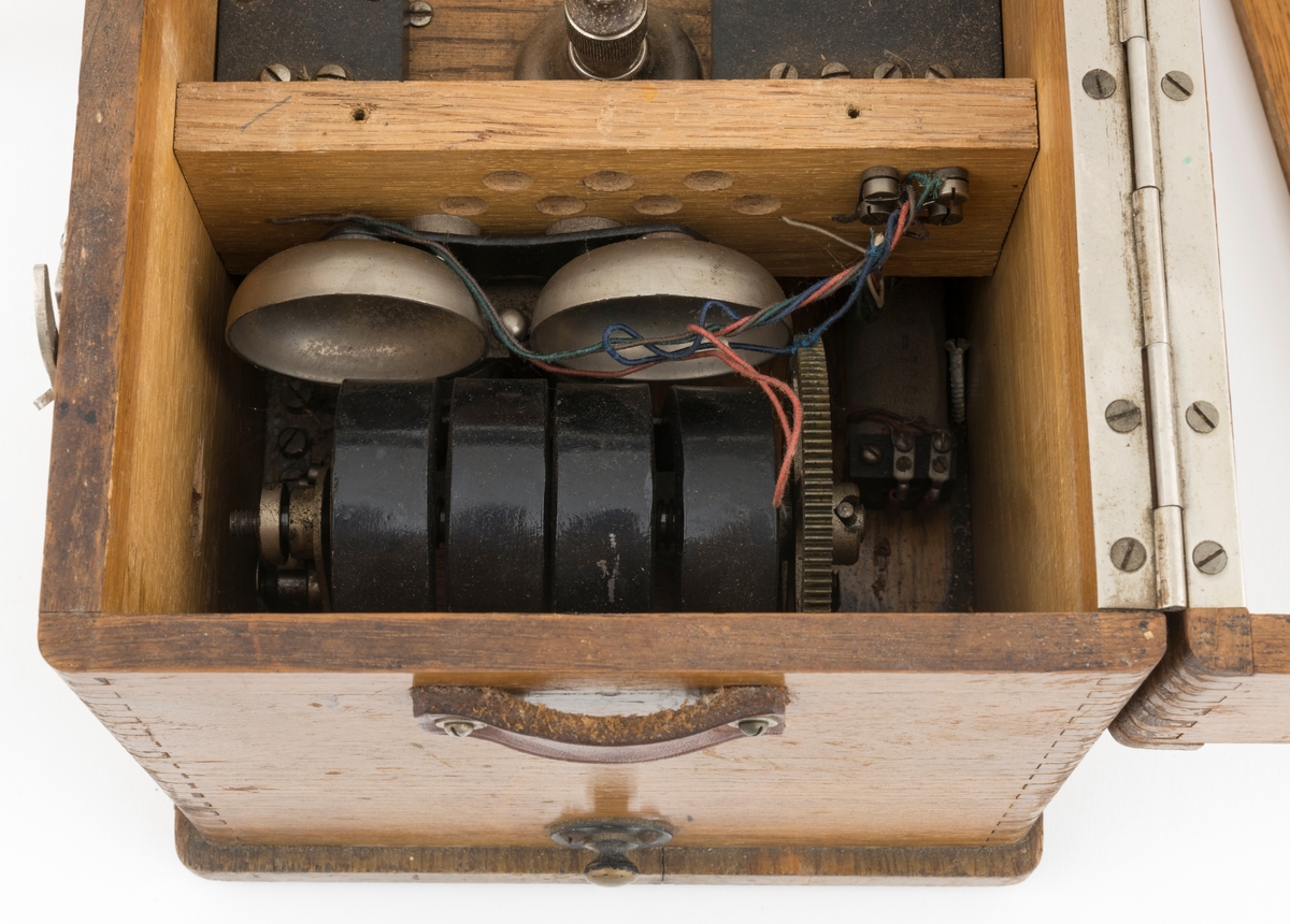 Bærbar felttelefon, antakelig av type som ble brukt tidlig på 1900-tallet. Telefonen er montert i ei trekasse, antakelig lagd av eikematerialer. Kassa har ei botnplate som er 33,1 centimeter lang, 20,2 centimeter bred og 1,5 centimeter tjukk. Ved endene av botnplatas underside er det 19,3 centimeter lange og 3,1 centimeter brede trelister som  gjenstanden sto på. Oppå den nevnte botnplata er det ei 32 centimeter lang, 19 centimeter bred og 16,6 centimeter høy kasse med fingerskjøtte, noe avrundete hjørner. Til denne kassas bakre langside er det hengslet et lokk av samme type materiale og med samme lengde og bredde som kassa. Lokket er 6,1 centimeter høyt. Lokket stenges ved hjelp av dreibare kroker på kassas frontside som vendes, slik at de omslutter skruer på lokkets frontside. Litt lengre neden på kassefronten er det et gjennombrutt hull med 1,3 centimeters diameter, antakelig innføringspunktet for sveiva til telefonen. Dette hukket kan stenges ved hjelp av ei lita, dreibar stålskive når sveiva ikke er i bruk. Da festes sveiva i en trekloss som er innlimt i et hjørne i lokket. Telefonen bæres i ei cirka 2,4 centimeter bred lysebrun lærrein, som er festet i messingknapper, sentralt på den nedre delen av kassas tverrender. At kassa bæres med den riktige sida opp er sikret ved hjelp av smøyestoler av lær høyere oppe på de samme endeflatene. Lengden på bærereima kan justeres ved hjelp av ei nålespenne. Inne i kassa er det antakelig et batteri, skjult under trelokk i to plan. På det nedre er det inntappet en toleddet, forkrommet holder for telefonrøret. Også telefonrøret er forkrommet, med har bakelittkomponenter for håndgrep (midten), øreanlegg og taletrakt. Sentralt på lokkets underside er det pålimt en trekloss med et tjukt svart filtbelegg, som skulle holde telefonrøret på plass under transport. Ellers er det innlimt to etiketter på undersida av lokket. Den ene er et firmamerke som refererer til «AKTIESELSKABET ELEKTISK BUREAU, KRISTIANIA, NORGE». På dette merket er det et rødt skjold med en løvefigur med krone over. Undersida av lokket er det også påklistrert en etikett med tekniske spesifikasjoner fra produsenten. Denne kontrollseddelen har også vært nummert, datert og signert, men hjørnet hvor blant annet årstallet har stått, er dessverre avrevet.