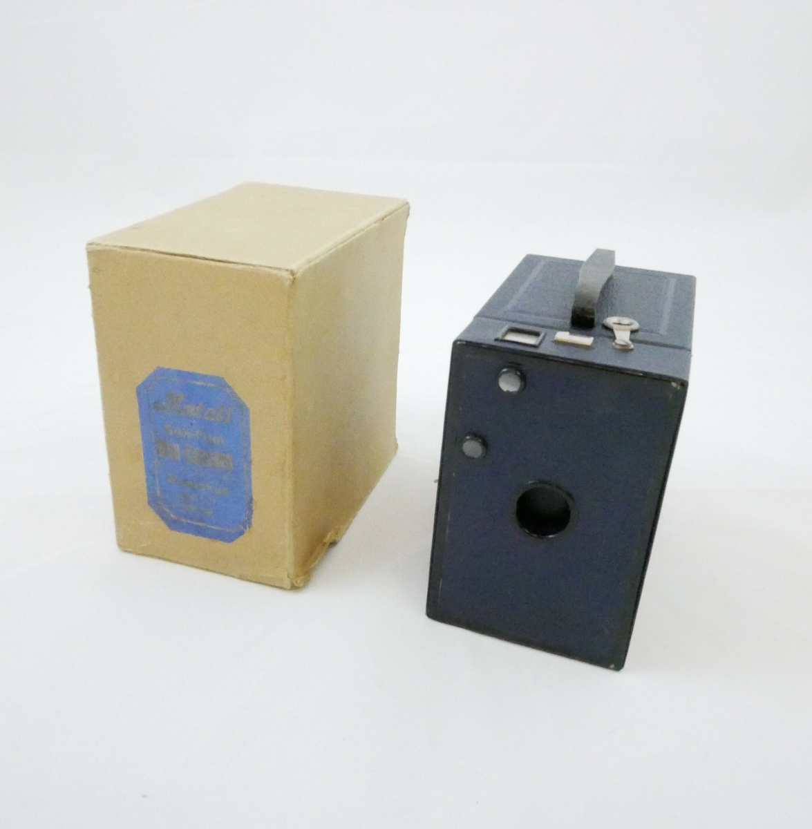 Brun pappemballasje som kan åpnes i den ene siden. Emballasjen er festet med stifter i den ene enden. På den ene kortsiden er det påklistret en merkelapp med blå bakgrunn og bokstaver i gullfarge.

Påskrift: 
Metall // Roll- Film // BOX- CAMERA // 6 ½ X 11 - 2 ½ X 4 ​​​1⁄4 // MENISKUS // Foc. 1 : 11 // Nr. 160

Inne i emballasjen er det et kamera. Kameraet er inne i et etui av blått lær, på toppen er det et svart håndtak i samme materiale. Etuiet kan åpnes i den ene siden og er festet med to metallhekter. 