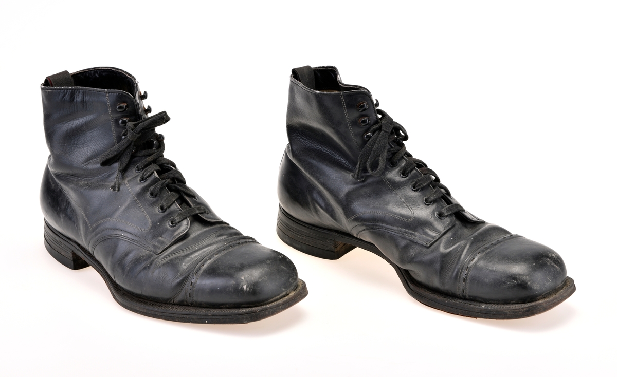 Et par svarte sko/støvler til mann. De er sydd sammen av tre deler av svart skinn. Tåen er svrundet firkantet. På Tåen er det en gjennomhullet skinnbord. Ytterste del av tåen er forsterket. I front er det snøring med 5 par metallmaljer og 4 par metallhekter. De er snøret med flate, svarte skolisser. Under snøringen er det en uforet skinntunge. Skoene er foret med hvitt lerretsstoff. Innersålen er av brunt skinn med forsterkning på hælområdet. Det er tekst på innersålen, men den er delvis slitt bort. Oppe bak på skoen er det en svart og rød lerretshempe sydd på. Skoene er randsydd. Yttersålen er av lær. På hælen er det satt på gummihæl med stift.