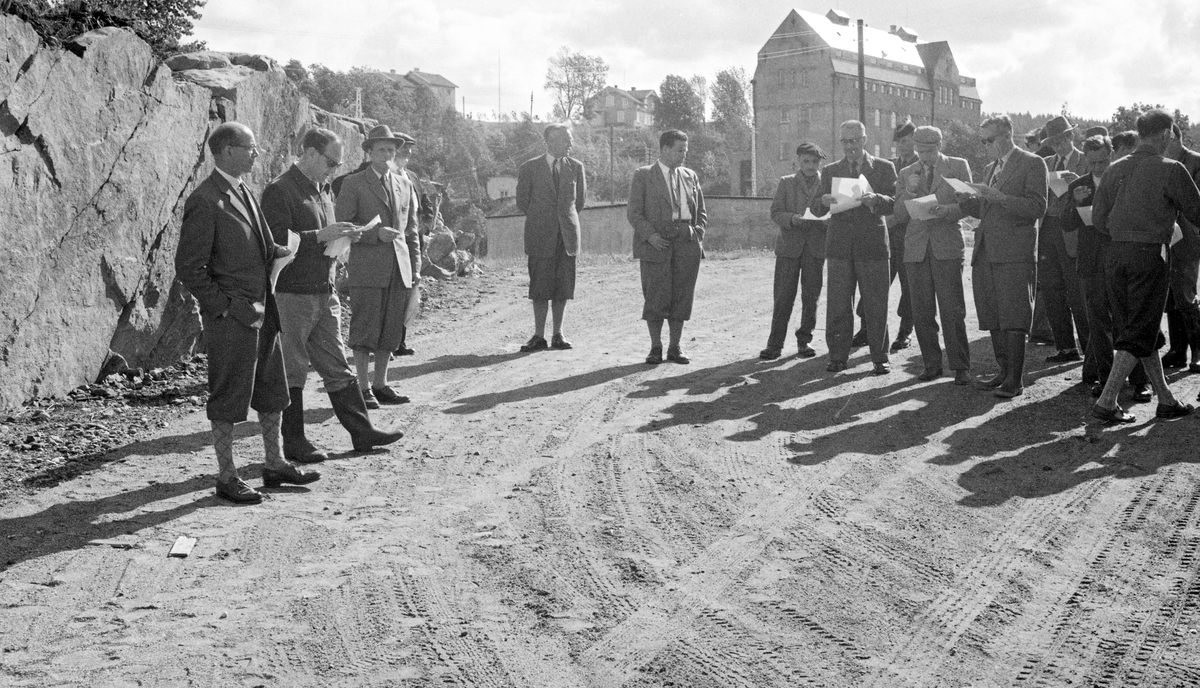 I juni 1959 hadde FN-organisasjonen FAO-ECE Commitee on Forest Working Techniques and Training of Forest Workers fagkonferanse i Norge. Konferansen samlet 60 deltakere fra 17 ulike land. Konferansen hadde også et ekskursjonsprogram med besøk hos skogbruksaktører på begge sider av Oslofjorden. Dette fotografiet skal være tatt ved Kykkelsrud i Askim, der representanter for Glomma fellesfløtingsforening redegjorde for hvordan de fløtet tømmer i et vassdrag der det også var flere store kraftverk. Her ser vi fløtingsdirektør Kaare Sparby og en av hans medarbeidere til venstre, mens professor Ivar Samset, som organiserte konferansen, tok seg en røyk. I bakgrunnen ser vi kraftverksbygningen.