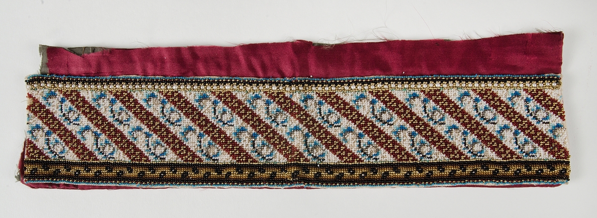 Fragment av klädesplagg med påsydd stramalj med pärlbroderi i pärlemor, guld, stål, rött, svart, vitt, blått, gult och brunt. Den pärlbroderade bården är fäst på vinrött siden, fodrad med grå taft.