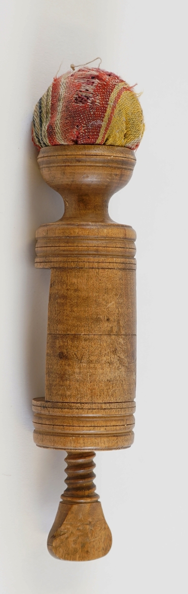 Ei syskrue i tre, med pute i tekstil på toppen. Nytta til å setje fast arbeidet til bordet når ein sydde.