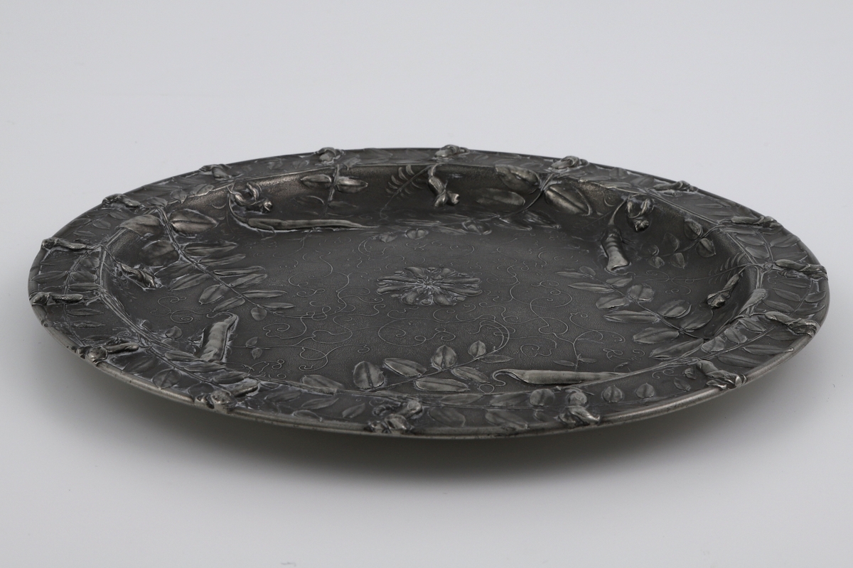 Fat i tinn. Langs den smale kanten løper plantestengler med små belger og blader. Inn mot fatets sentrum skyter tidvis større belger, bladverk og plantespirer, og midten er markert med en rosett. Dekoren skal forestille erteblomstplanten vesce des haies (fr. gjerdevikke), og er utført i relieff.