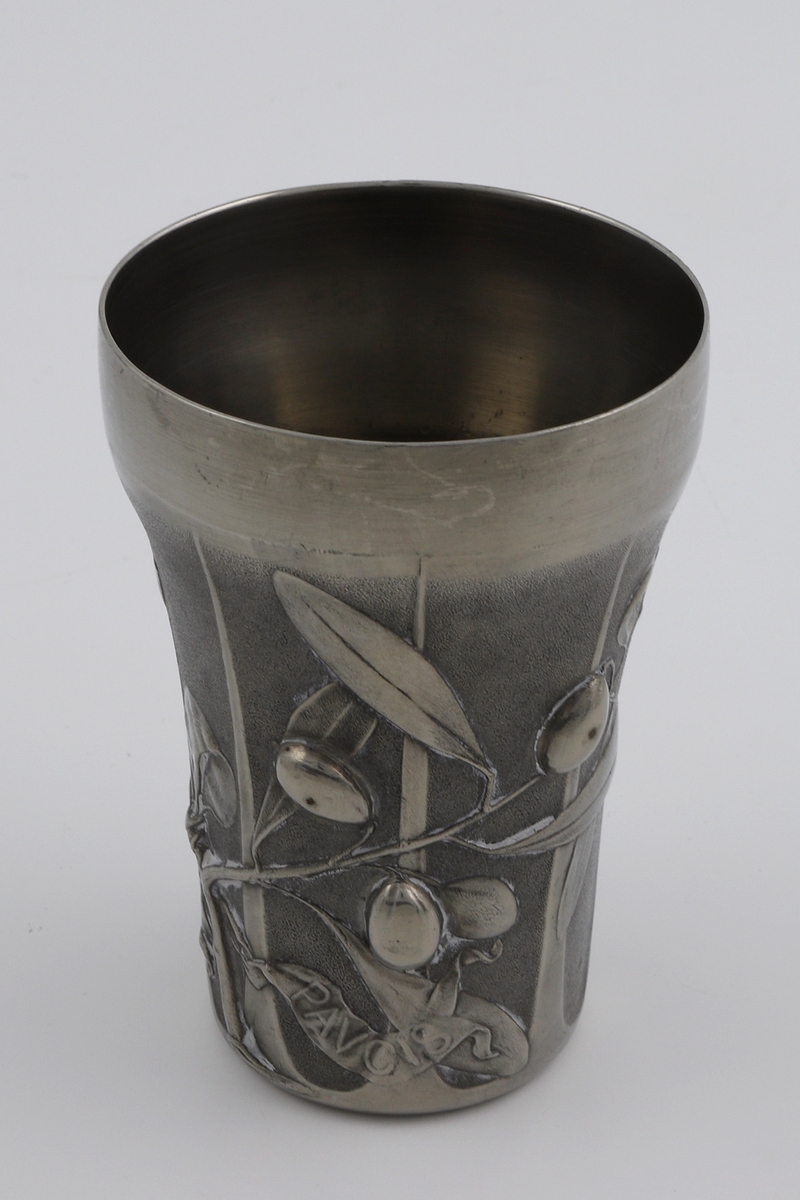 Beger i tinn. Formen smalner svakt nedover. Dekoren på korpus viser en olivenkvist i lavt relieff, derav begerets tittel L’Olivier (fr. oliventre). Til grenen er det festet to bånd med innskriften «MVLTA PAVCIS» (lat. mye med lite). Kanten er blank og glatt, mens korpus trolig er patinert. Yttersiden er inndelt med gjentagende rifler.