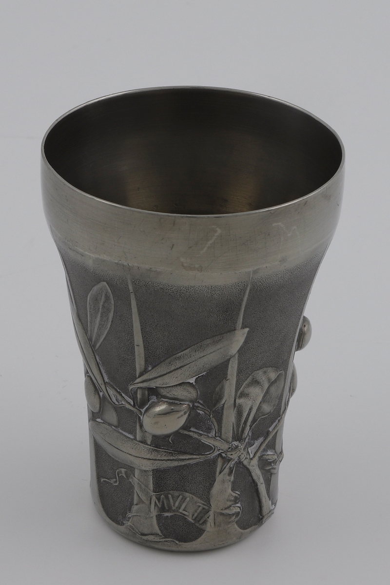 Beger i tinn. Formen smalner svakt nedover. Dekoren på korpus viser en olivenkvist i lavt relieff, derav begerets tittel L’Olivier (fr. oliventre). Til grenen er det festet to bånd med innskriften «MVLTA PAVCIS» (lat. mye med lite). Kanten er blank og glatt, mens korpus trolig er patinert. Yttersiden er inndelt med gjentagende rifler.