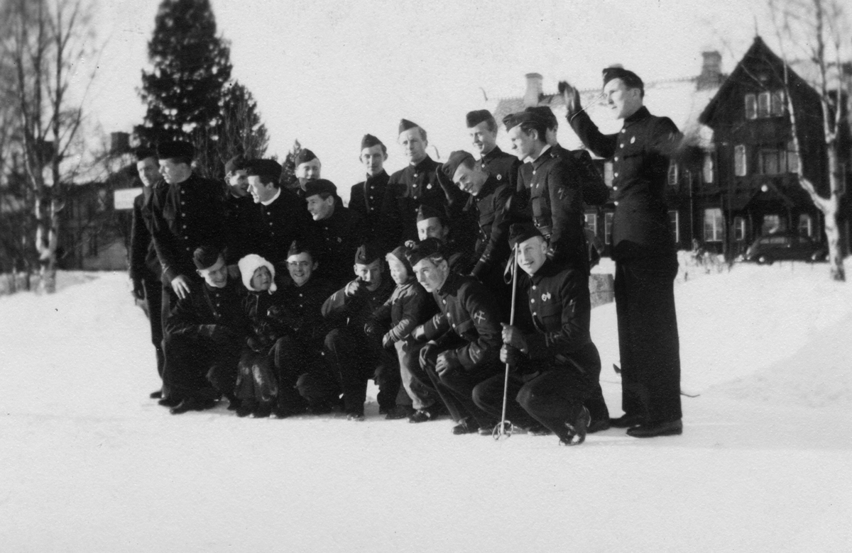 Grupporträtt av deltagare i korpralskola på F 7, tidigt 1940-tal. Militärer och två barn samlade utomhus i snön.