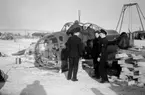 Flygtekniker och flygvapenpersonal samlade vid bärgat flygplan B 18 på kajen i Härnösands hamn efter nödlandning på isen den 10 februari 1946. Motorerna har demonterats. I bakgrunden syns en lyftkran som användes vid bärgningen.